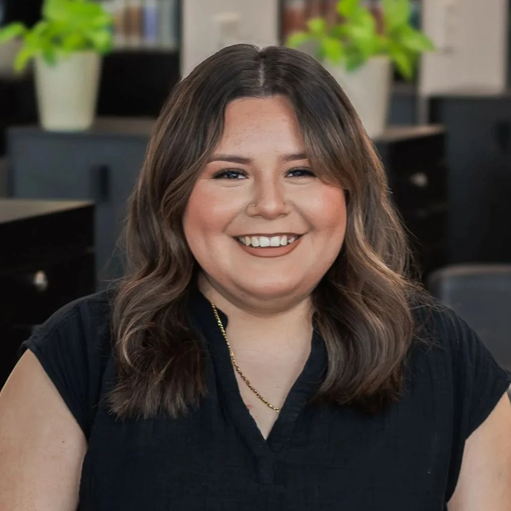 A woman with shoulder-length brown hair smiling in an office setting with black desks and potted green plants in the background.