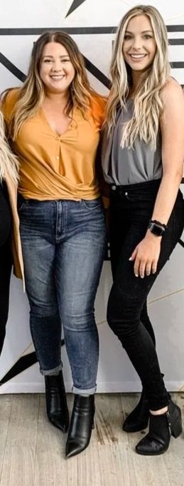 Four women standing together in front of a modern geometric wall design with a black and white logo. They are smiling and dressed in casual, stylish clothes.