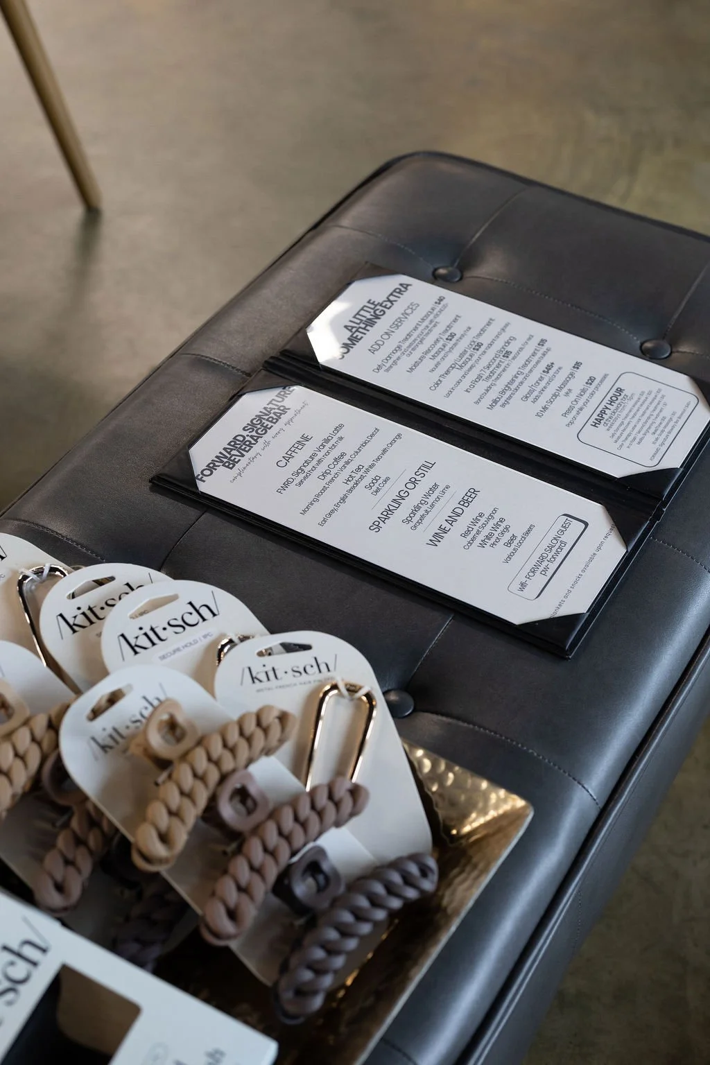 Menus on a black leather padded bench with beige woven hair ties branded 'kit-sch' in the foreground.