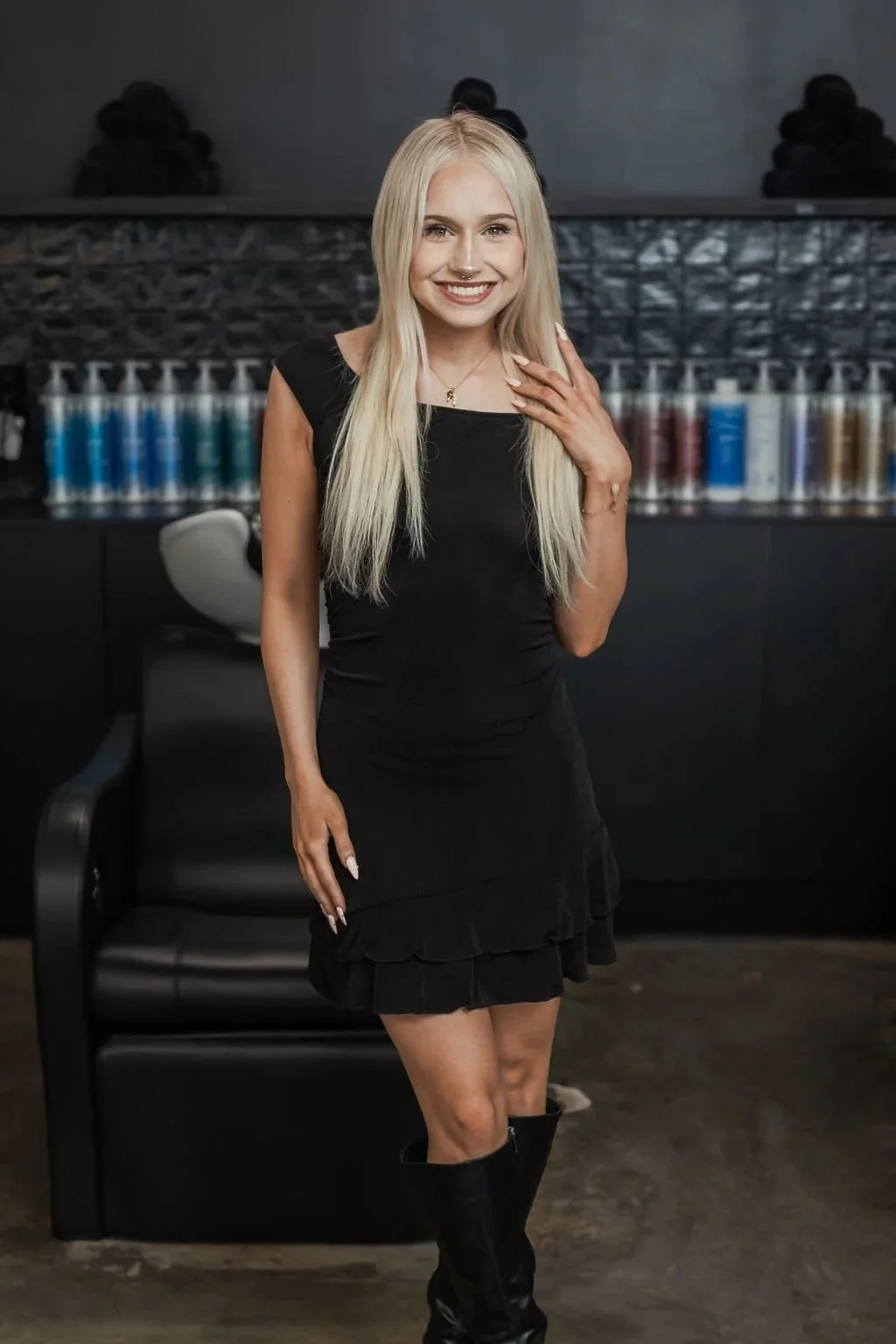 A young woman with long blonde hair and light skin, wearing a black dress and black knee-high boots, smiling in a modern salon or studio setting with hair products on shelves in the background.