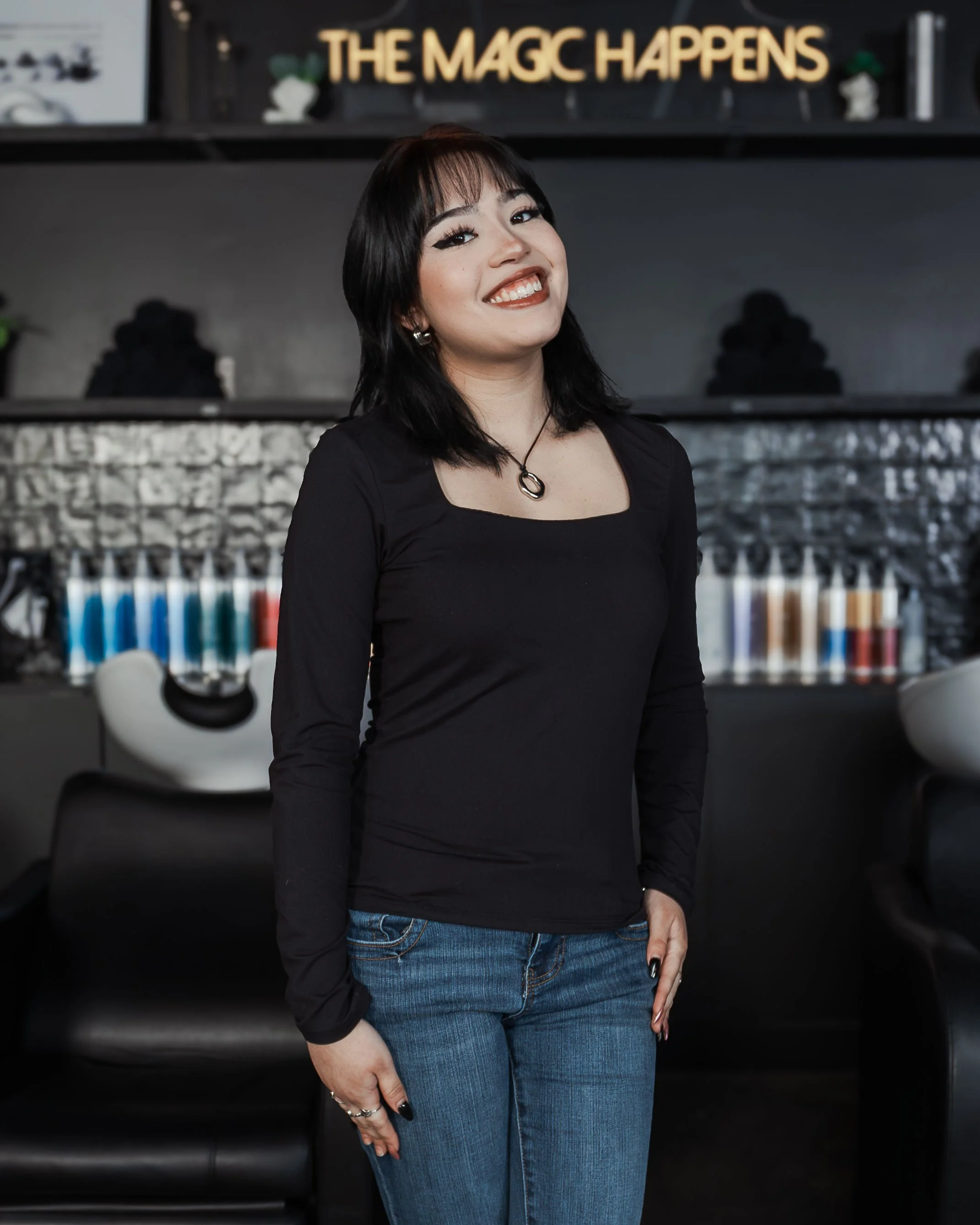 A woman with dark hair, wearing a black top and blue jeans, smiling in front of a black shelf with a sign that says "The Magic Happens."