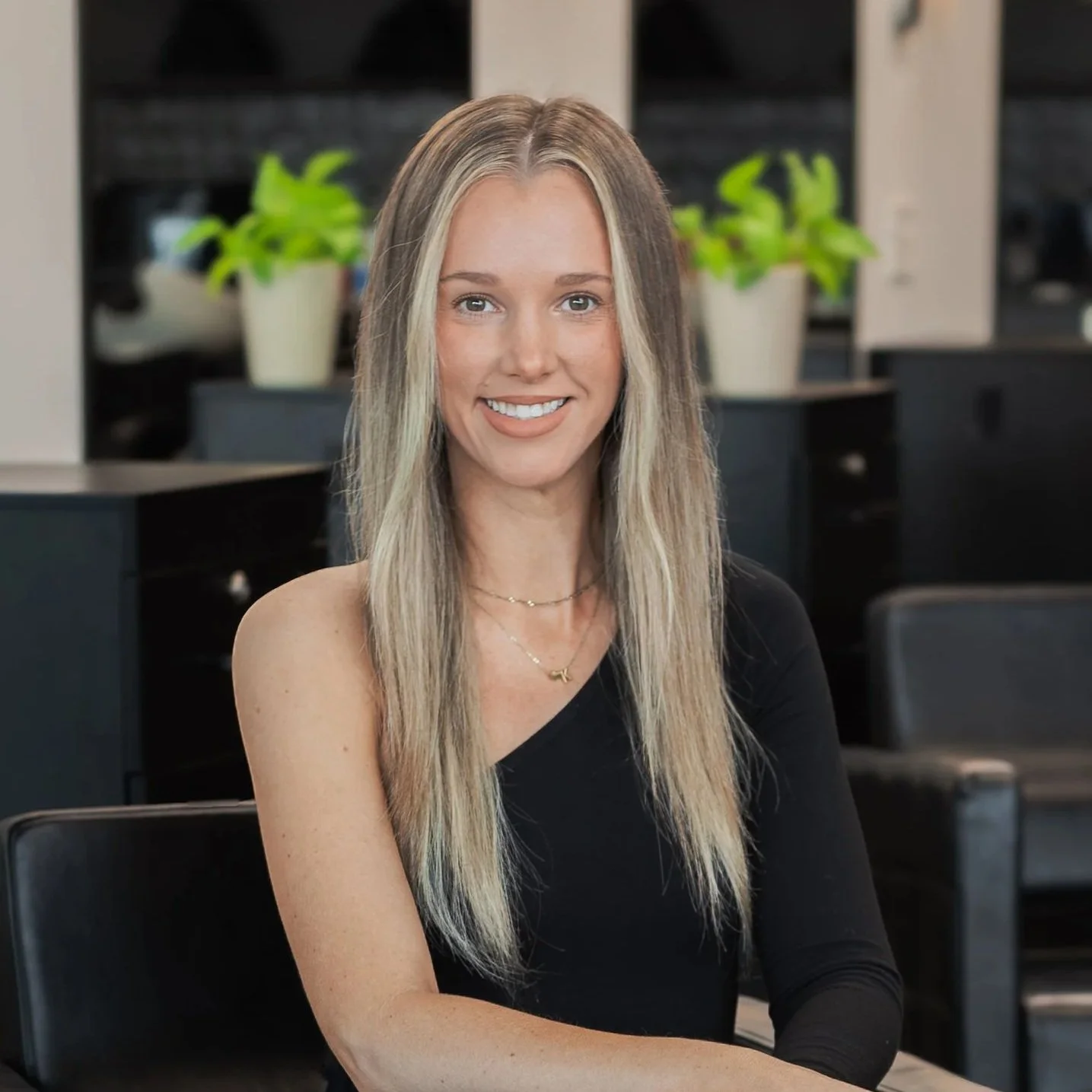 A young woman with long blonde hair wearing a black top, smiling, seated in an indoor space with plants and black furniture in the background.