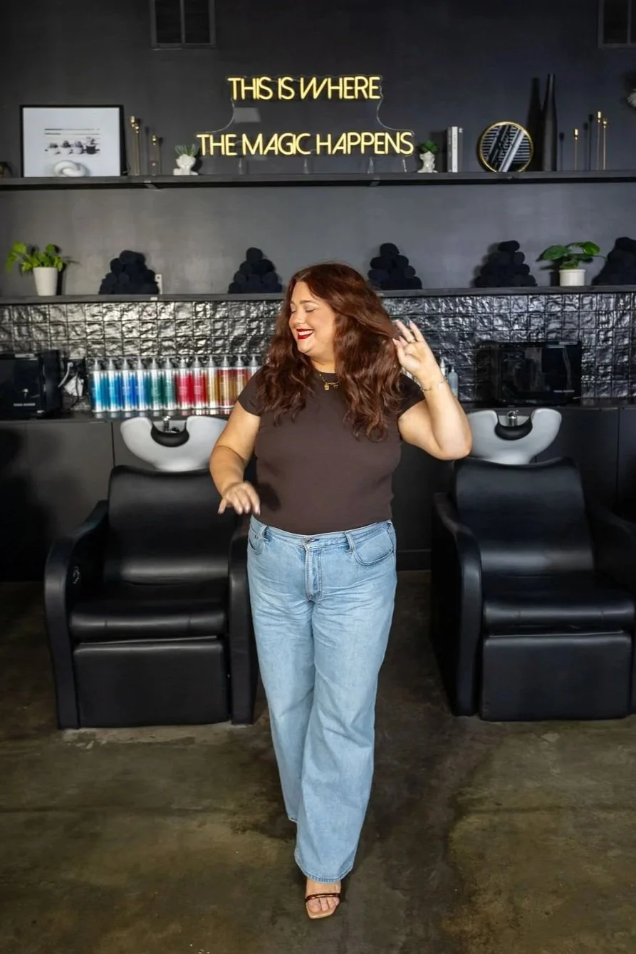 A woman with curly red hair, wearing a dark t-shirt and jeans, smiling and dancing in a salon with black chairs and a decorative black and gray wall behind her. There is a neon sign above her that reads "This is where the magic happens".