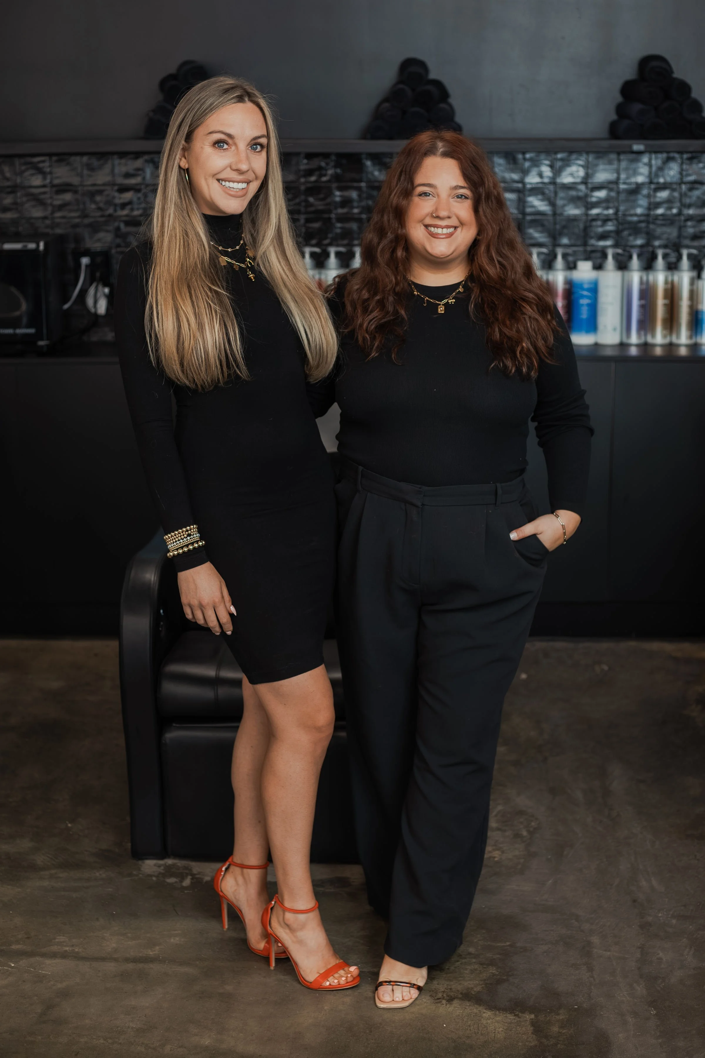 Two women standing side by side indoors, smiling at the camera. The woman on the left has long blonde hair, wears a black dress and orange high heels. The woman on the right has long curly red hair, wears a black top and black pants with open-toed shoes. The background includes a black shelf with hair products and black towels.