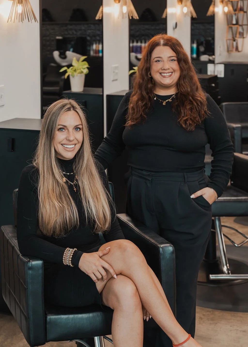 Two women with long hair, one seated with crossed legs, and the other standing beside her, both smiling and dressed in black, in a salon or stylist's workspace.