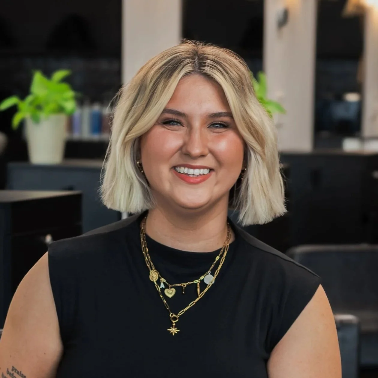 A smiling woman with shoulder-length blonde hair wearing a black sleeveless top and layered gold necklaces, sitting in a modern office environment with black furniture and green plants in the background.