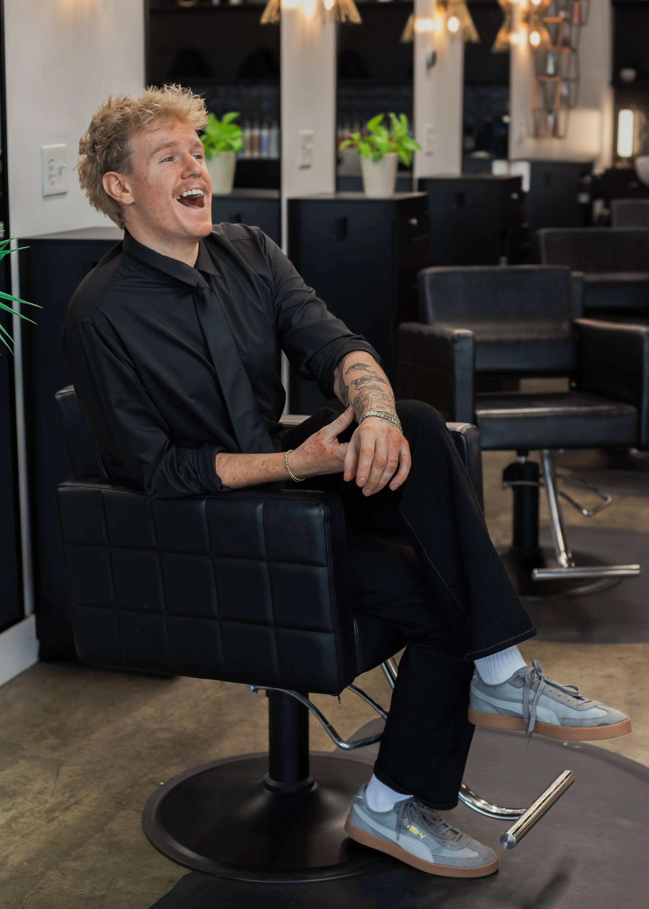 A young man with curly blond hair, tattoos on one arm, and wearing gray sneakers, sits in a black salon chair in a modern hair salon. He is smiling and appears to be laughing, with his mouth open and head slightly tilted back, in a relaxed and cheerful mood.