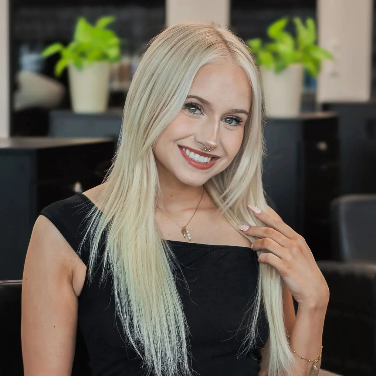 A young blonde woman with blue eyes smiling, wearing a black top and jewelry, indoors with green plants in the background.