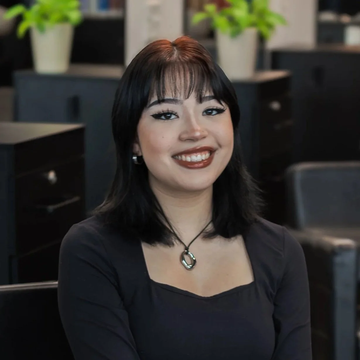 A woman with shoulder-length black hair and bangs, smiling, wearing a black top and a necklace, sitting in a modern office environment with black furniture and green plants in the background.