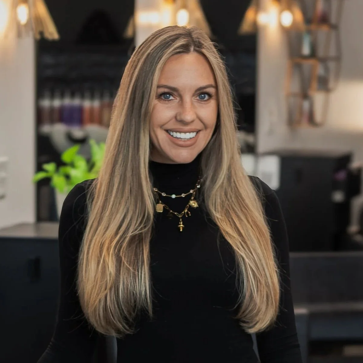 A smiling woman with long blonde hair wearing a black top and layered necklaces, standing indoors with blurred background including shelving and lighting.