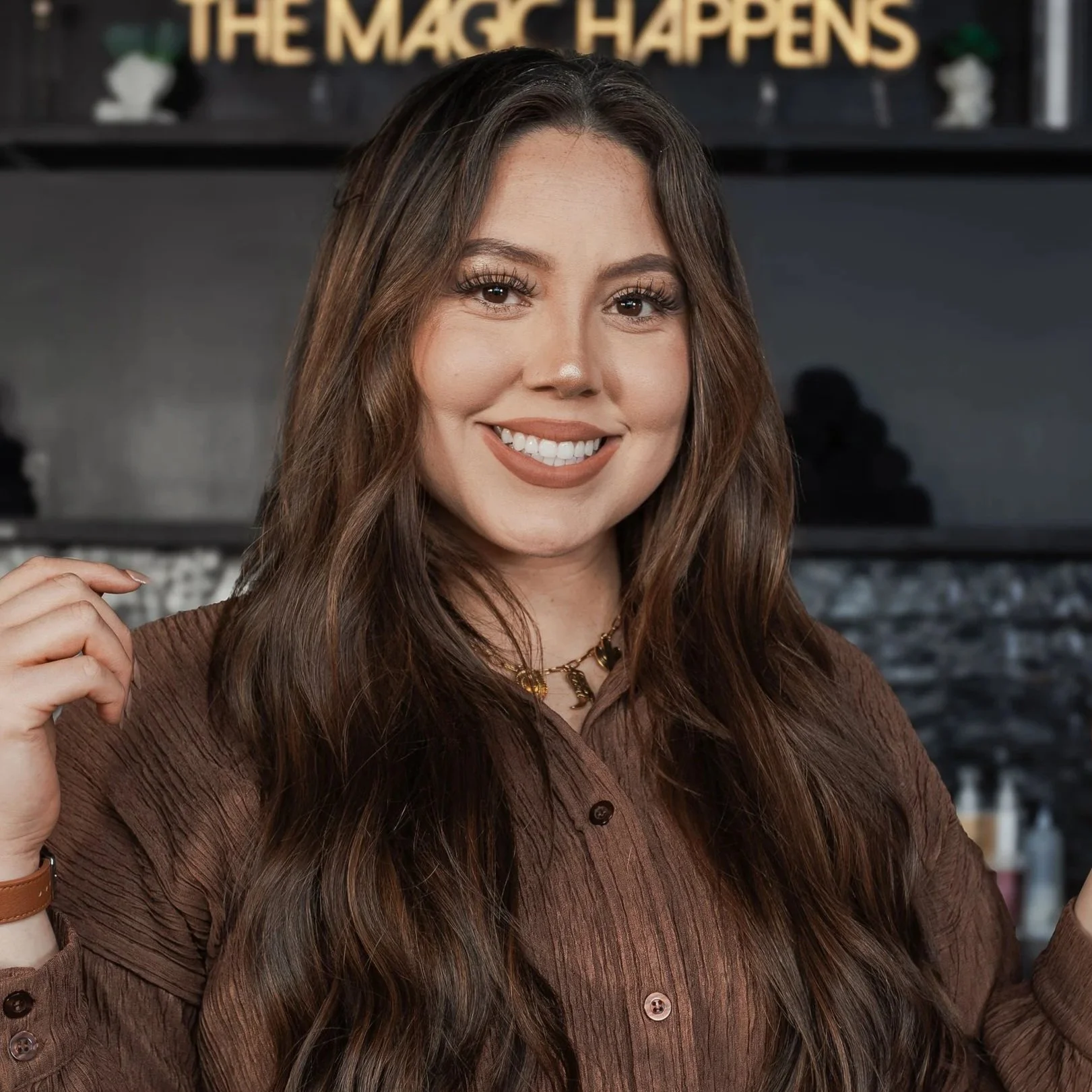 A smiling woman with long, wavy brown hair, wearing a brown shirt and gold jewelry, standing in front of a background featuring a sign that says 'THE MAGIC HAPPENS'.