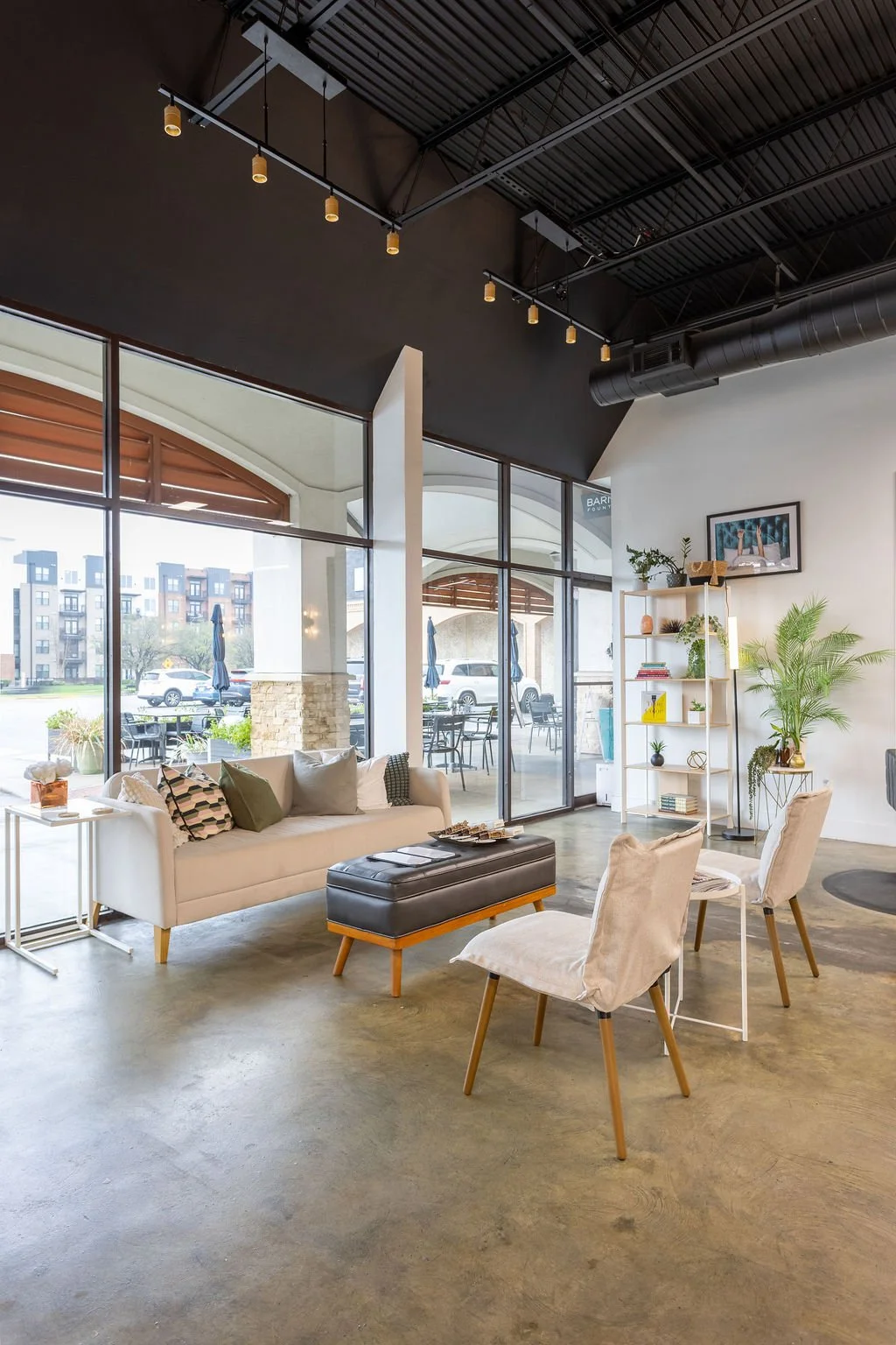 Modern lounge with white sofa, beige armchairs, black ottoman, large windows, and indoor plants.