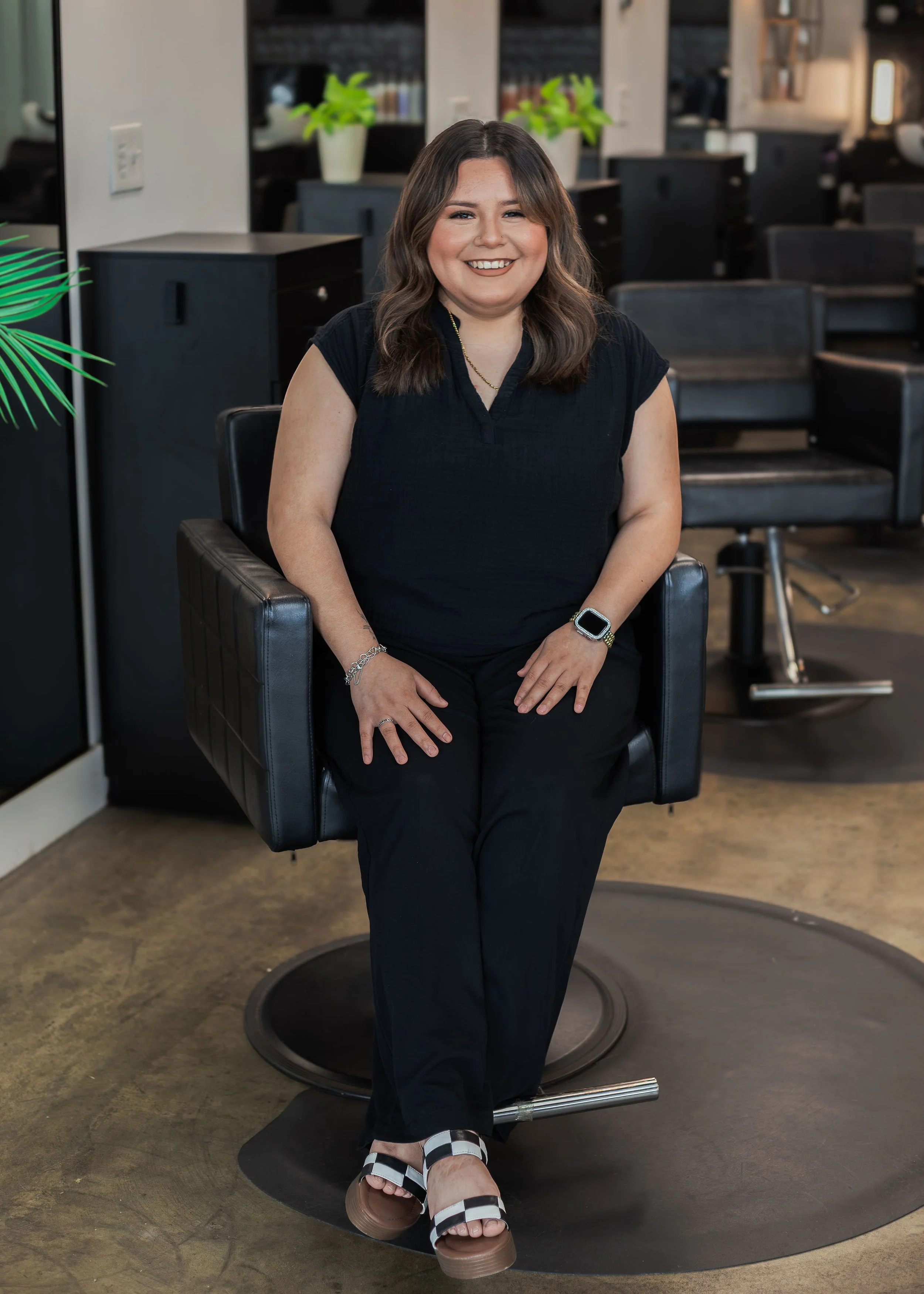 A smiling woman with shoulder-length wavy brown hair, sitting in a salon chair with black and tan checkered sandals, wearing a black top and black pants, with an Apple Watch on her left wrist, in a hair salon with black chairs, shelves with books, and potted plants in the background.