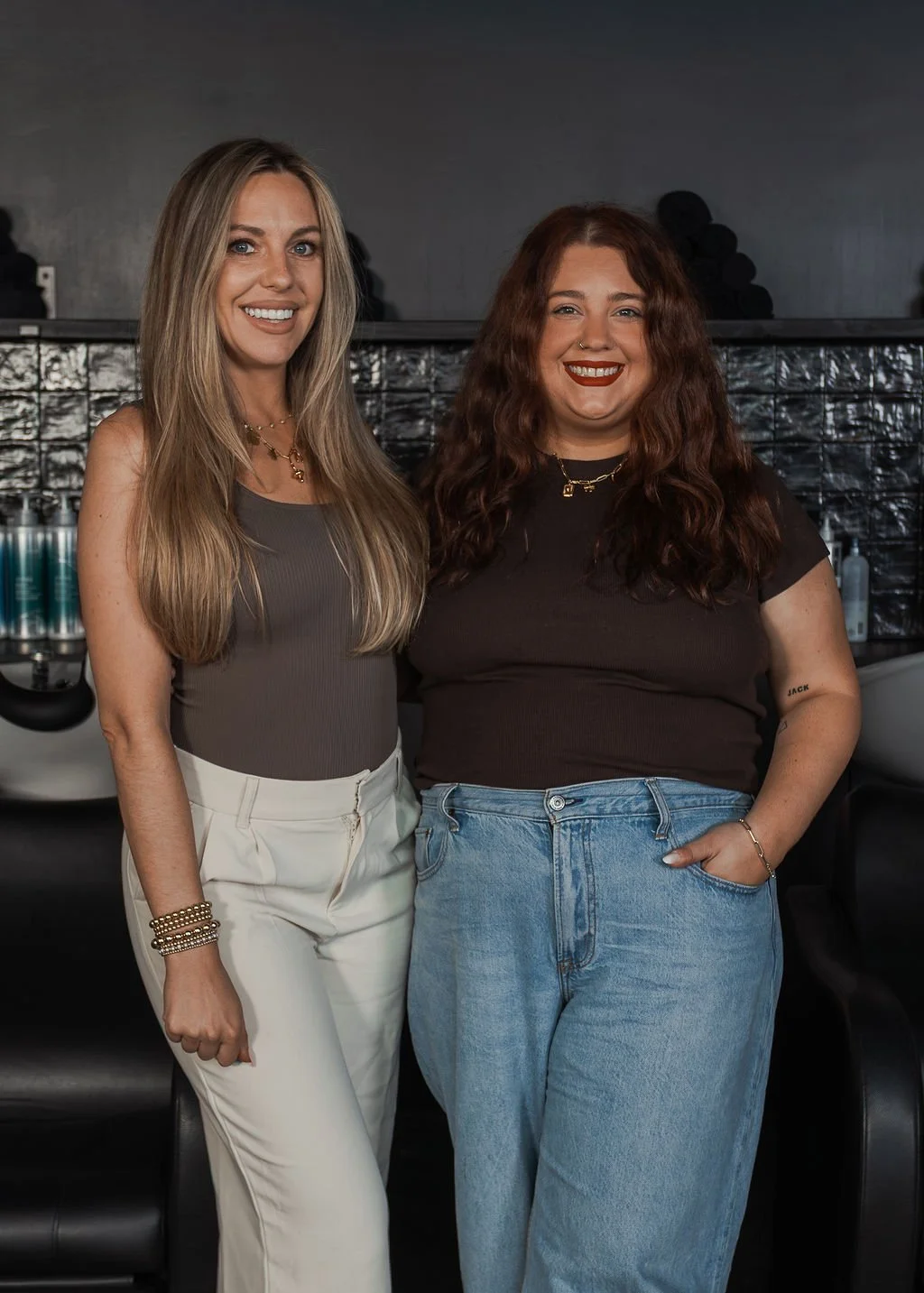 Two women standing close together, smiling for the camera in a modern, dark interior space.