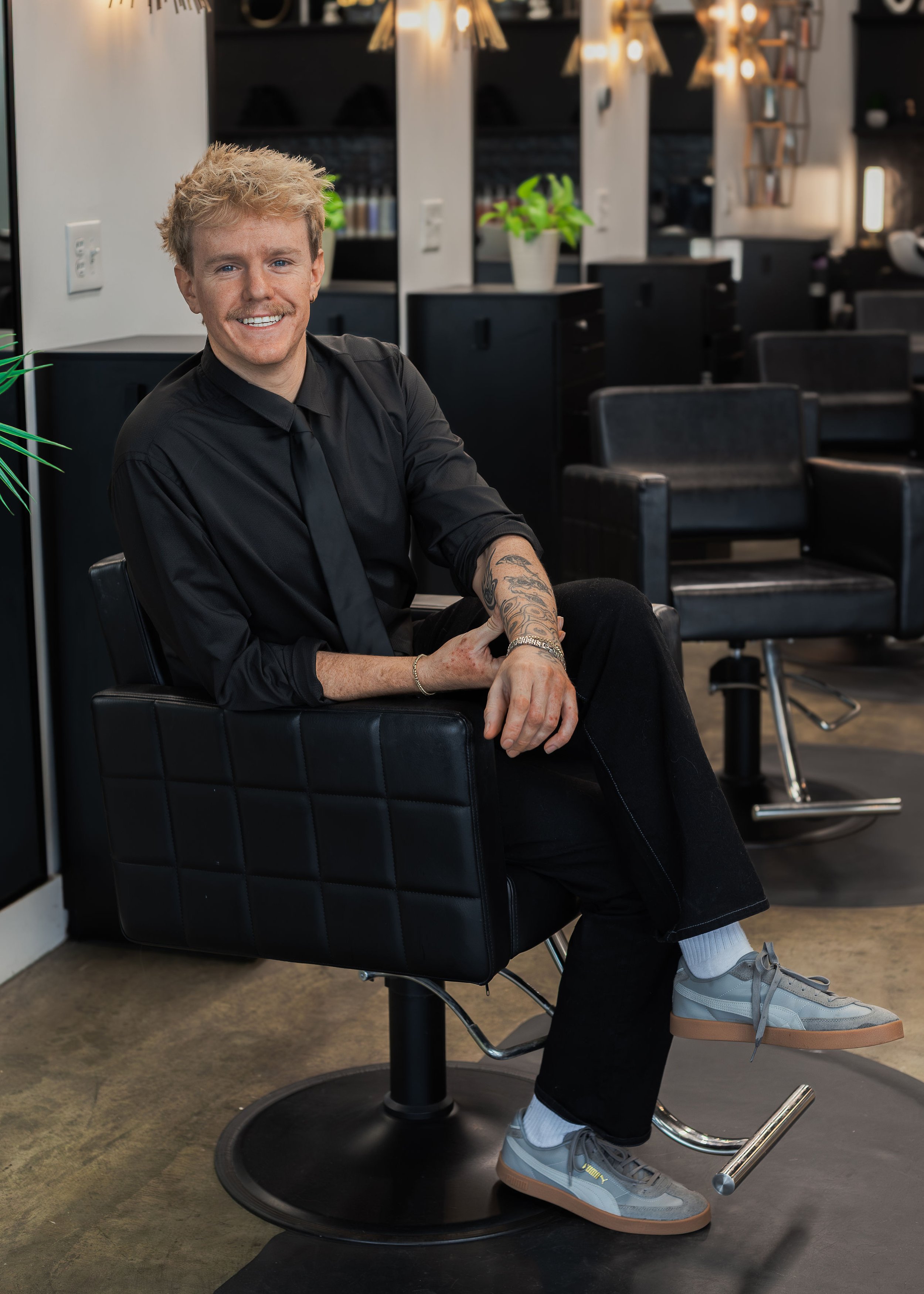 A young man with blond hair and a tattoo on his arm, dressed in black, sitting in a salon chair with a smile, in a modern hair salon with black furniture, plants, and warm lighting.