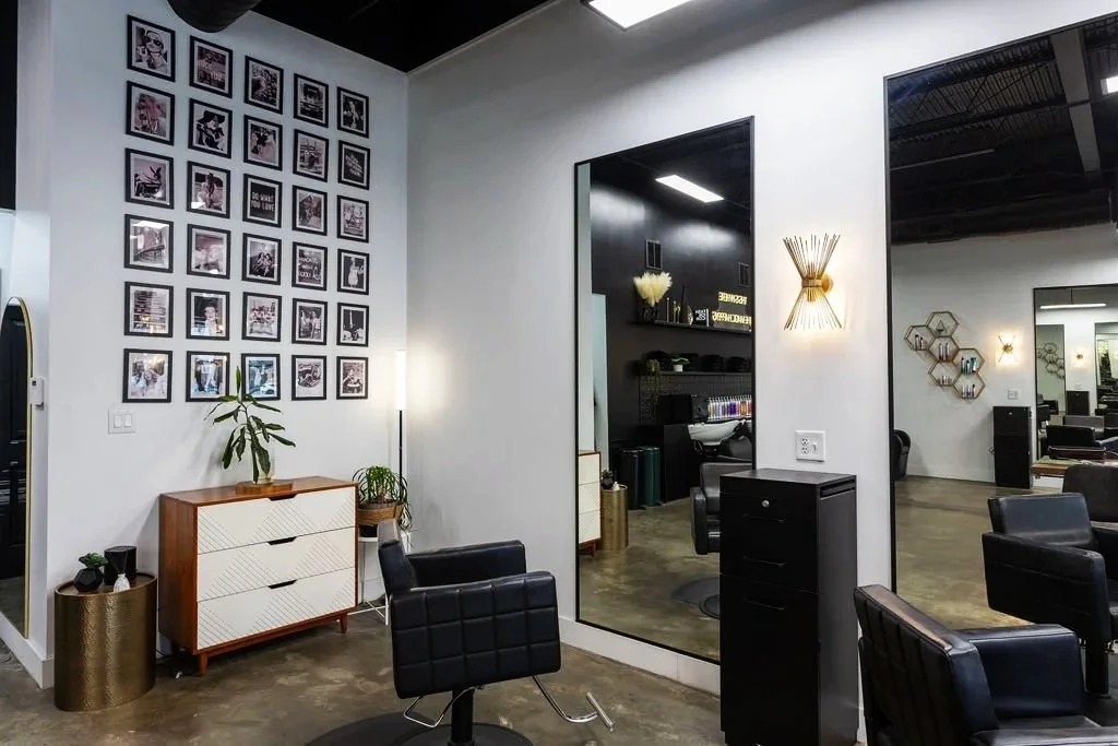 Modern hair salon interior featuring black chairs, a framed photo collage on the left wall, a white cabinet, plants, and decorative wall art.