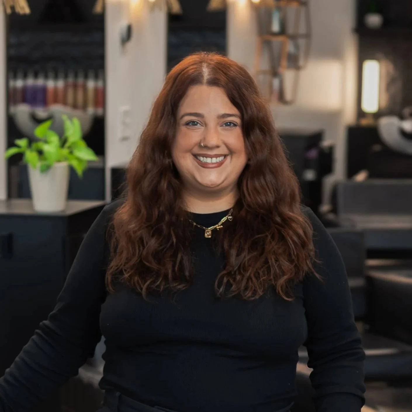 A smiling woman with long, wavy auburn hair, wearing a black top and a gold necklace, is in an office environment with black desks, shelves, and a potted plant in the background.