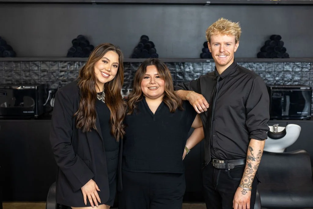 Three people standing together in a modern salon or salon-like setting, smiling at the camera.