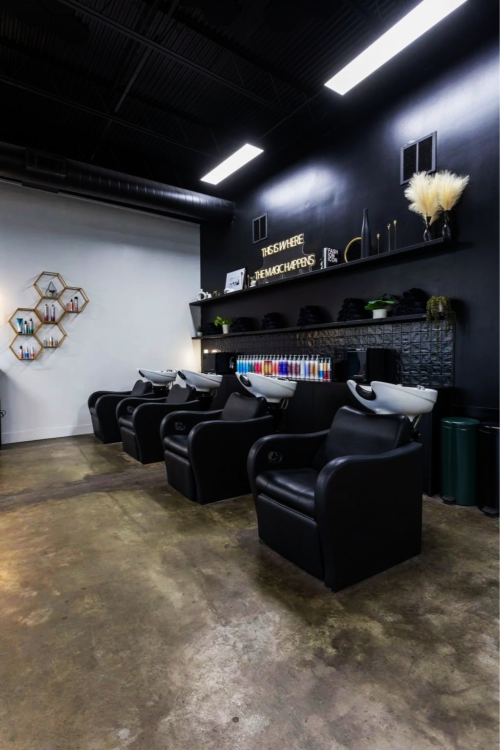 Hair salon with three black reclining chairs with white sinks, shelves with hair dye bottles, and decorative black and white accents.