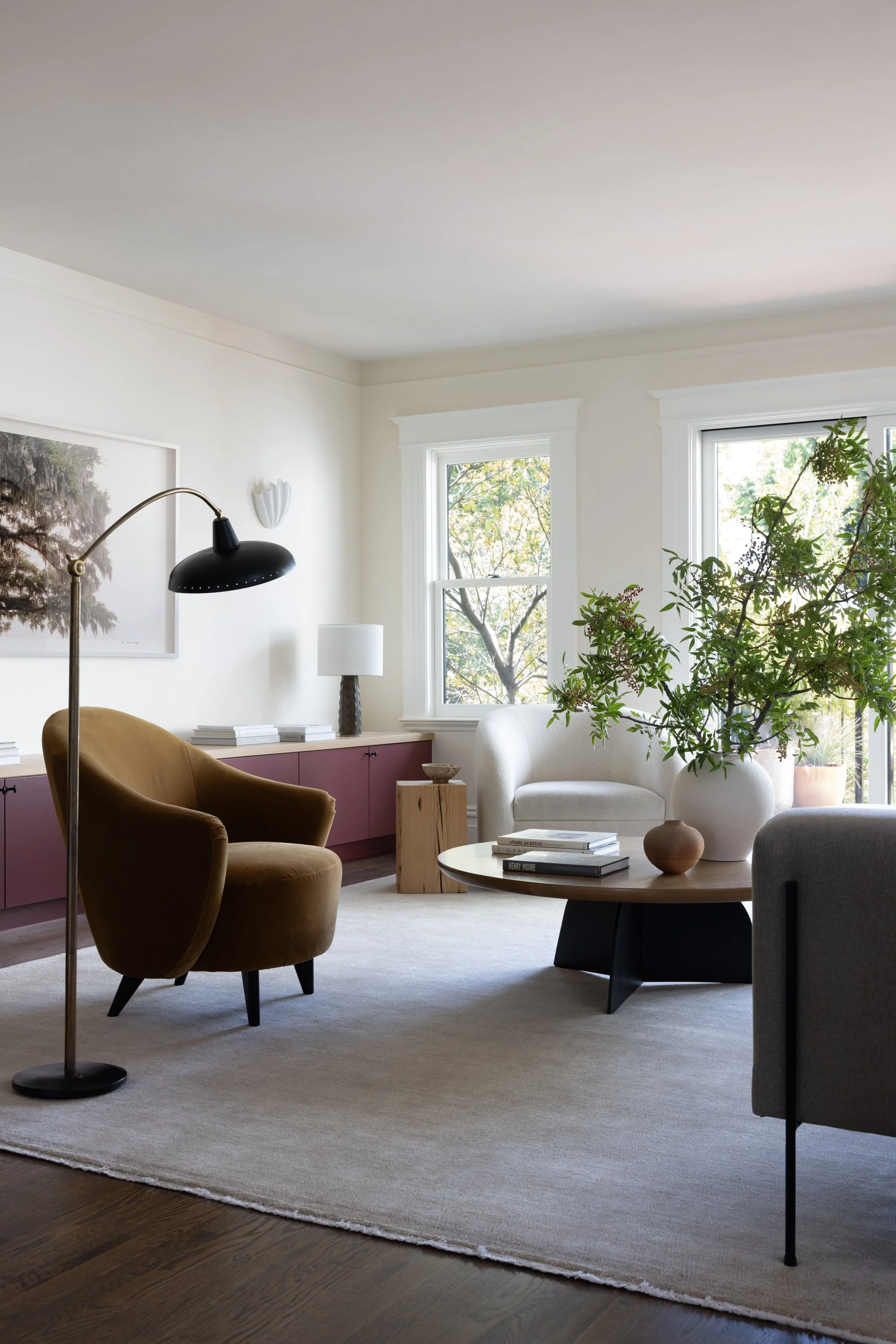 View of a different living room, showing side chairs, floor lamp, large round coffee table with vase and branches. A long sideboard can be seen with a table lamp, stacks of books, as well as large artwork on the wall, a wall sconce and large windows.