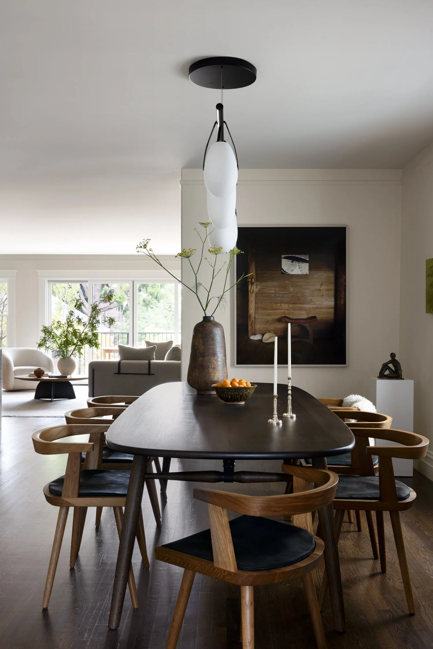 View of the dining table with chairs, large vase with flowers, bowl with small citrus, long candle sticks and candle holders, artwork visible in the back.