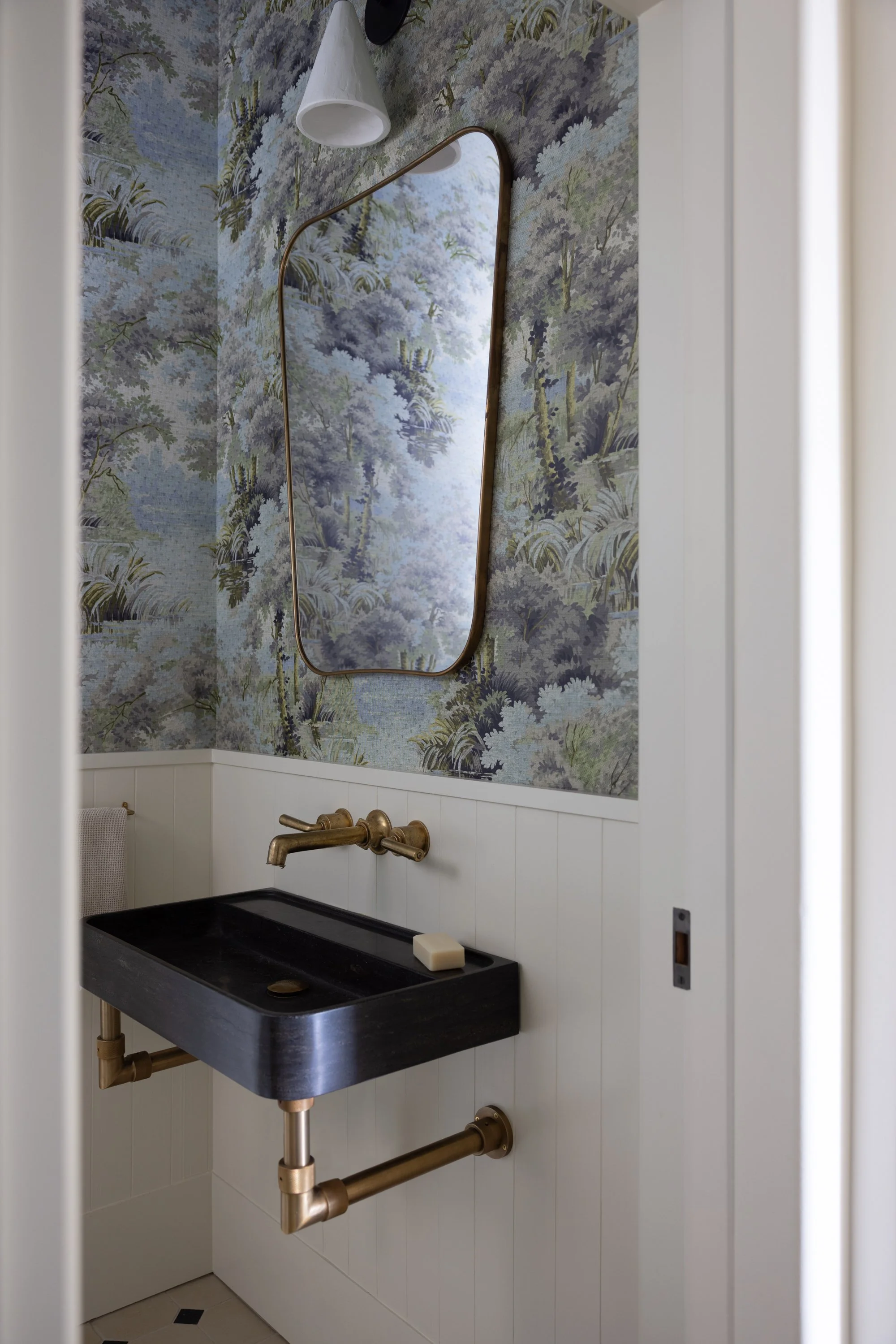 Vignette of powder room, with nature patterned wallpaper, large mirror, black sink and brass faucet.