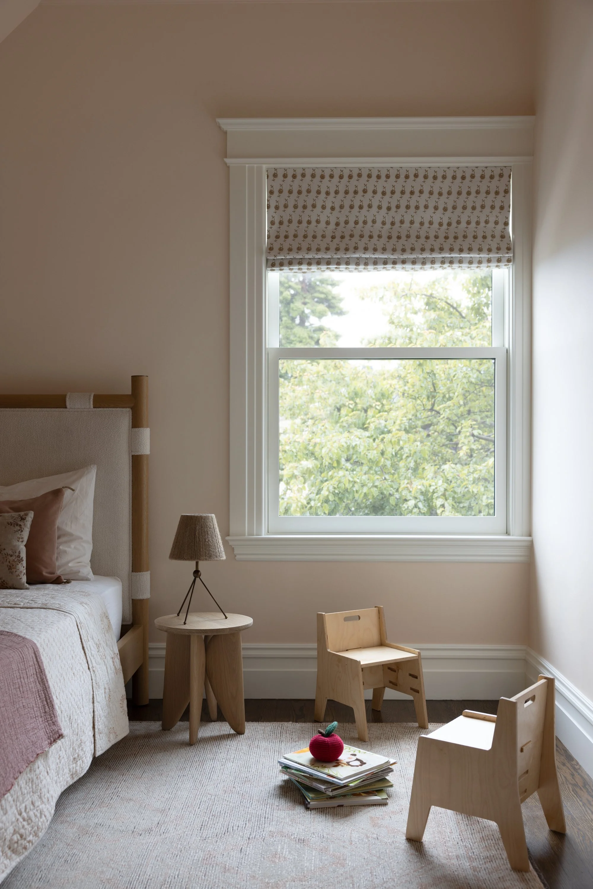 Children's bedroom with light rose colored walls, showing the bed, side table with table lamp and small wooden chairs and children's books.