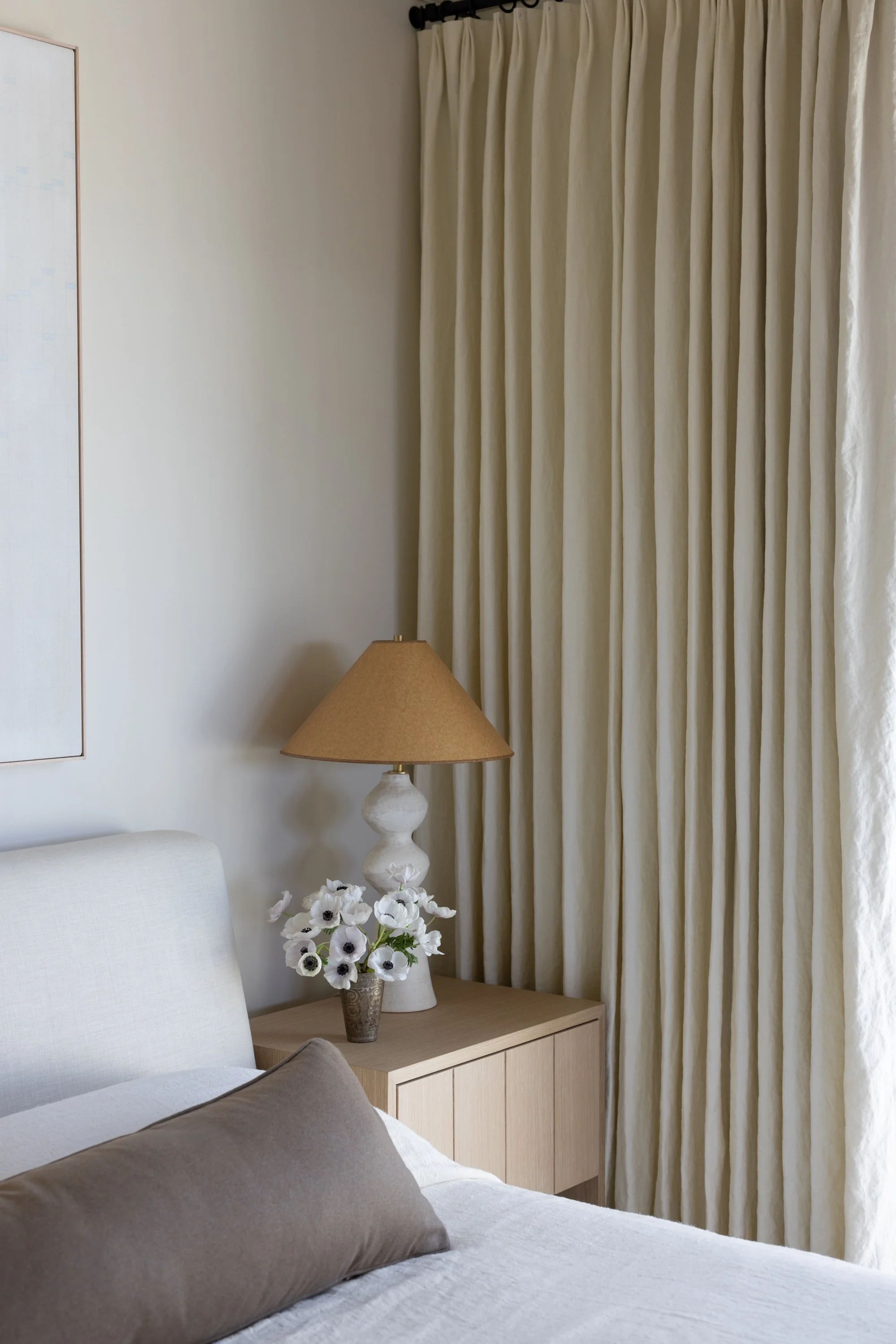 Closeup view of the light wood bedroom nightstand, showing a table lamp and vase with flowers and cream colored curtain in the background.