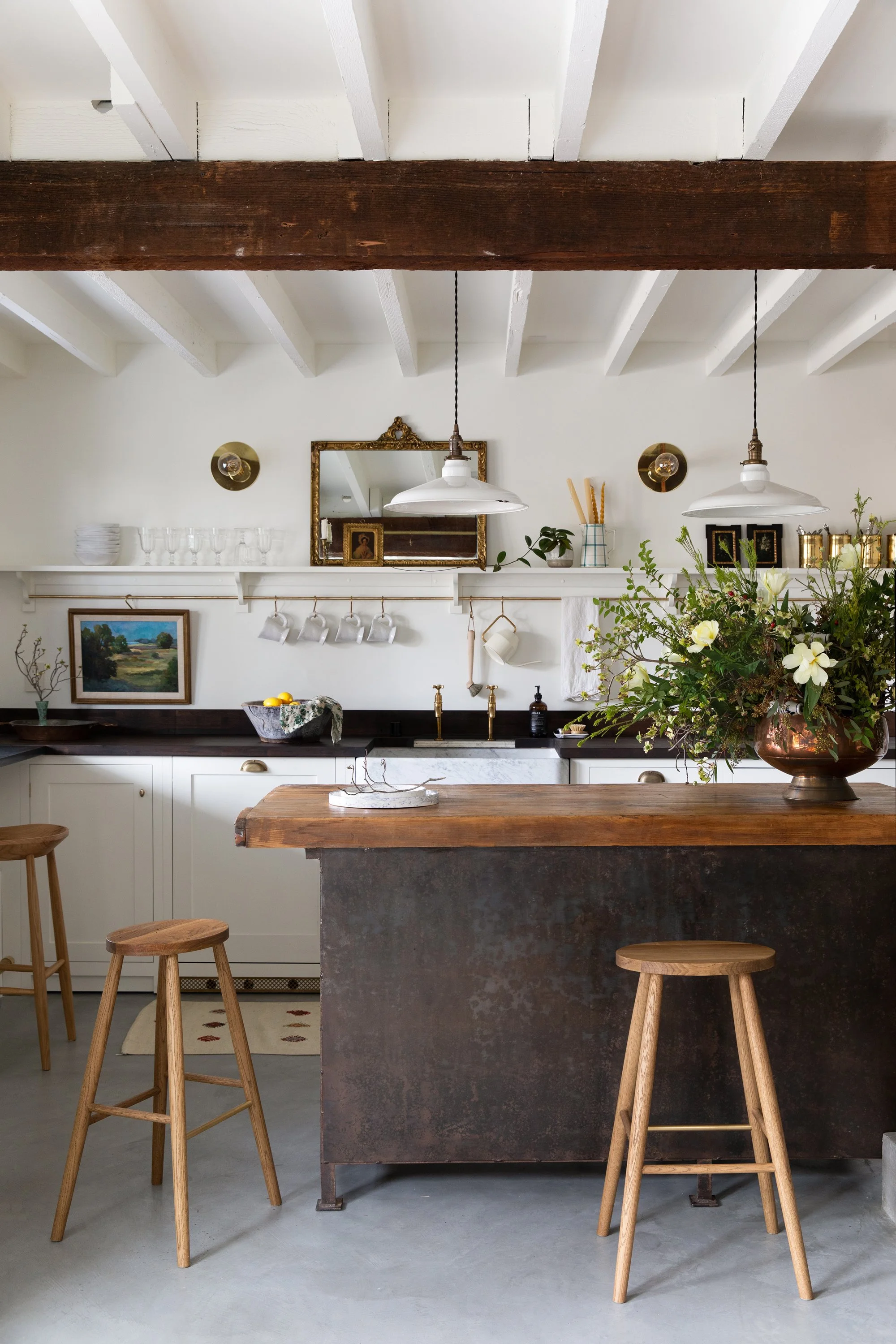 View of the kitchen, showing exposed beams, kitchen island, wooden stools, vase with large flower arrangement, lower cabinets with counters, sink, shelving with mirror, cups, glasses and bowls.