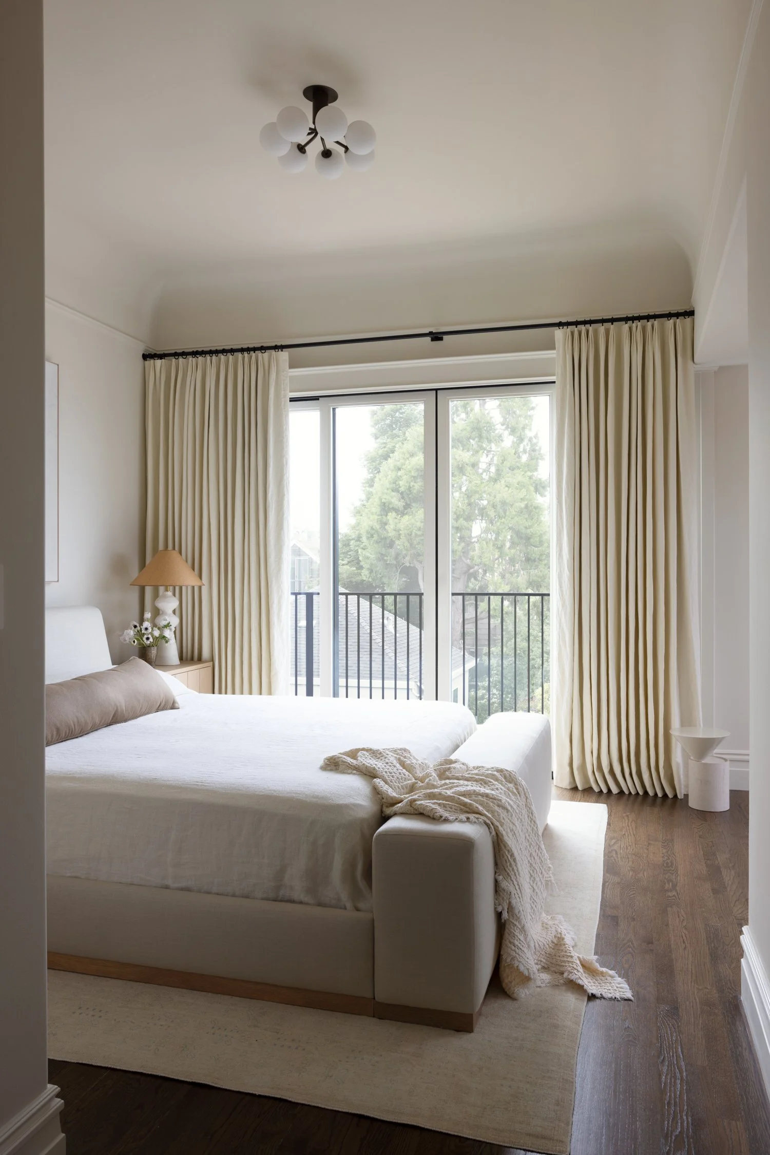 Side view of the bedroom, showing white and cream linens on a cream colored bed, with cream colored curtains.