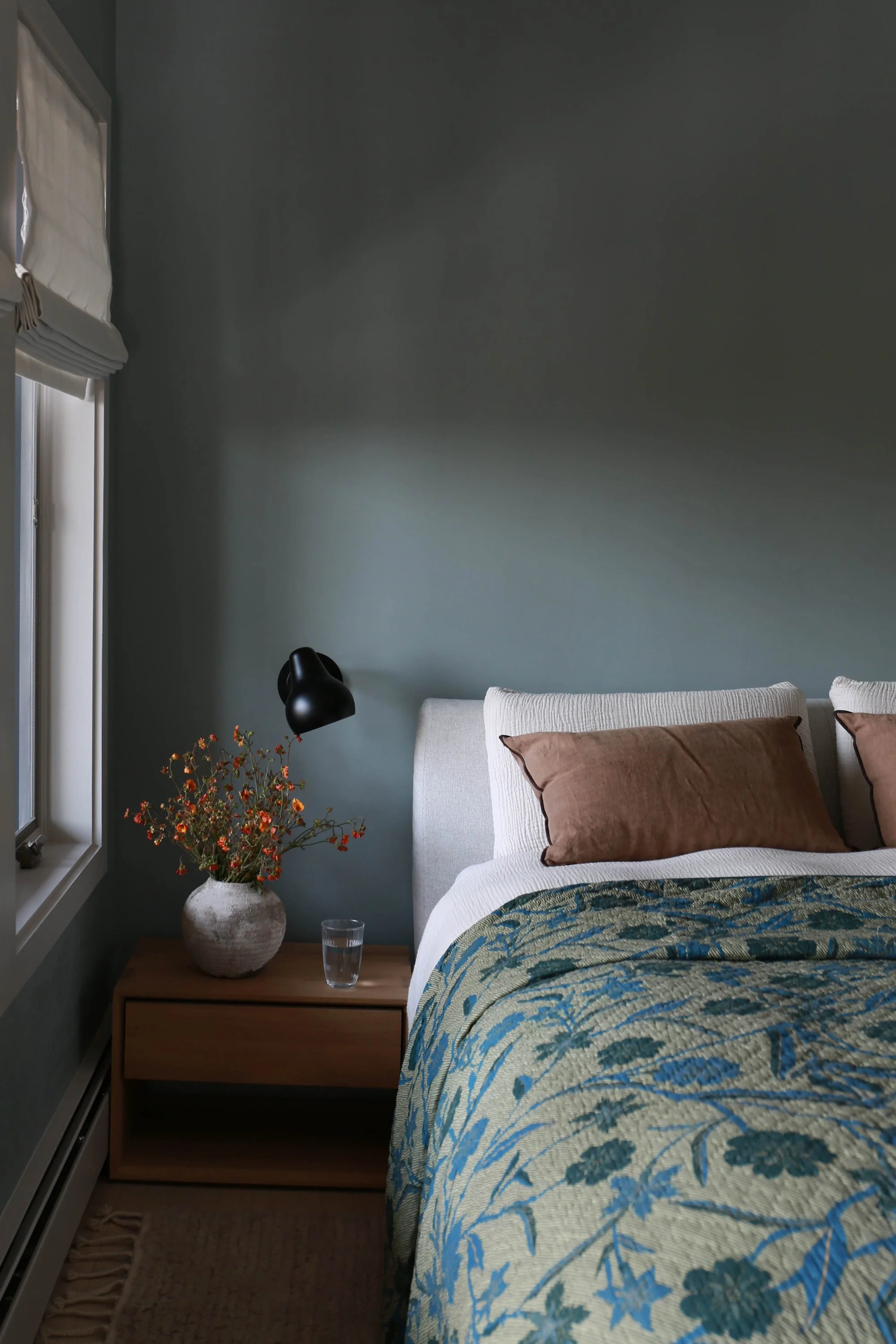 Bedroom view showing a flower patterned throw blanket, nightstand with vase and flowers.