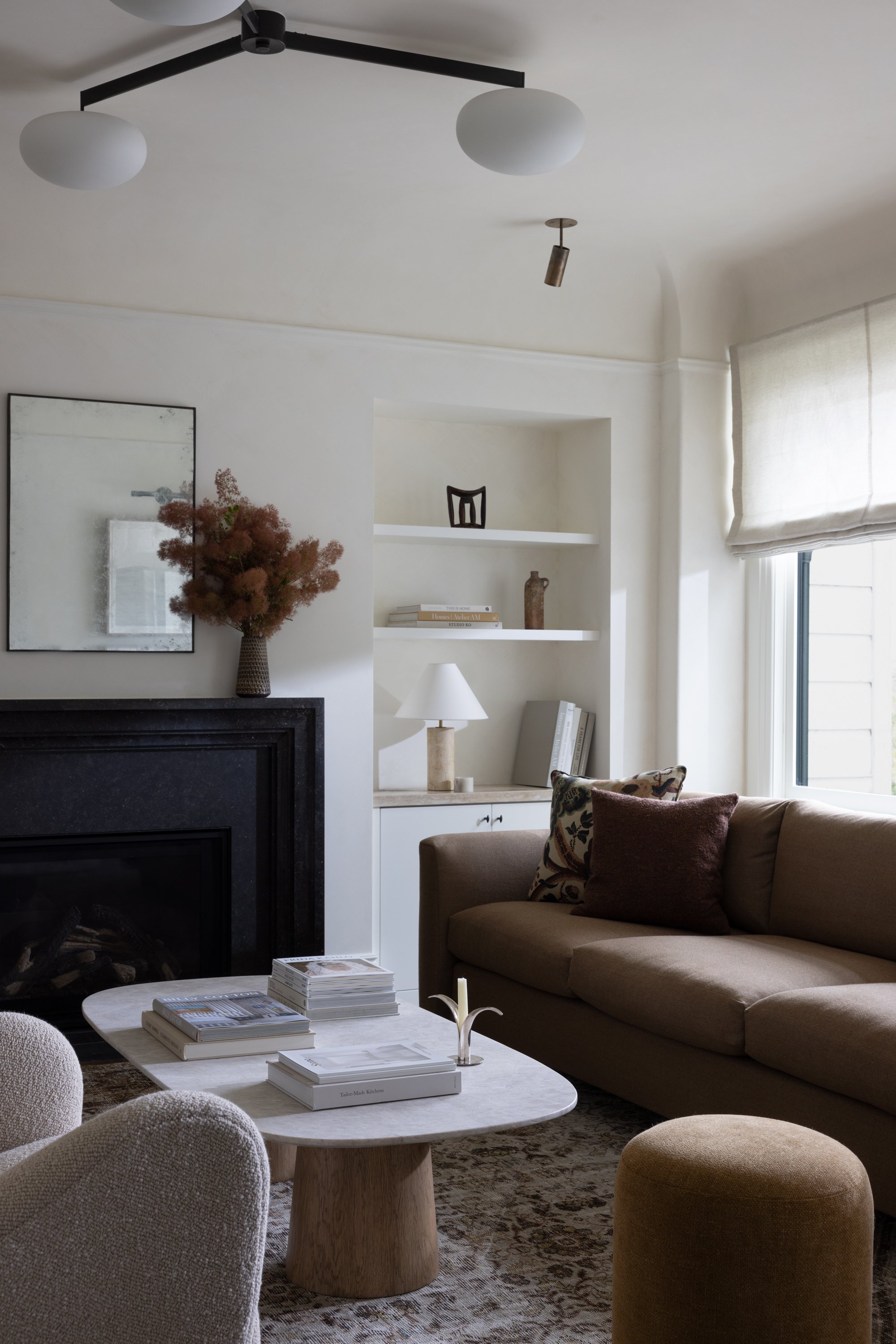 Living room side view showing a fireplace, a mirror, a vase with flowers, a sofa, side chair and pouf, recessed shelving with books, vases, art and table lamp, coffee table book with stacks of books, candle with candle holder, and decorative pillows.
