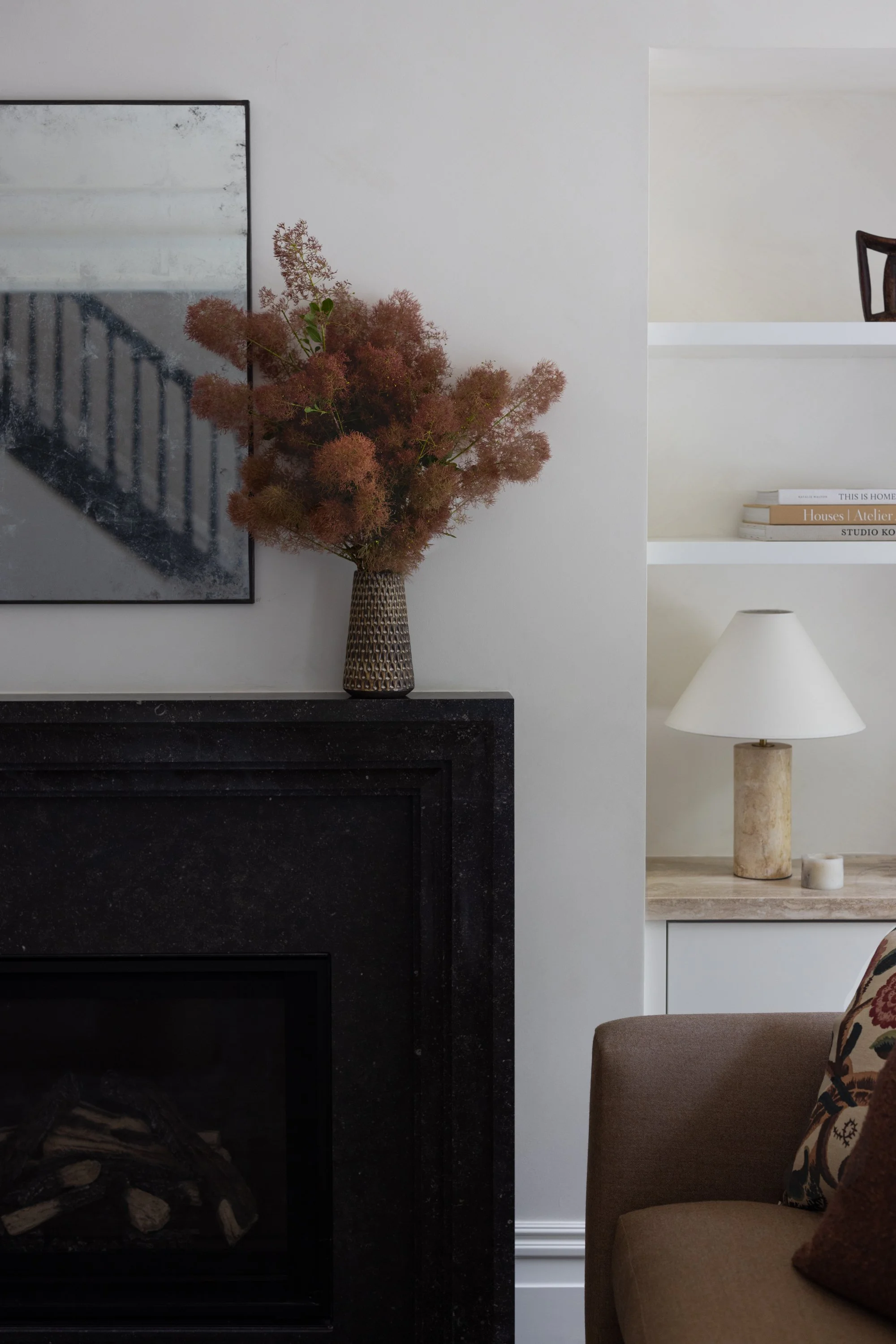 Closeup of fireplace in the living room, showing a vase with flowers, a mirror, recessed shelving with books, table lamp and art.