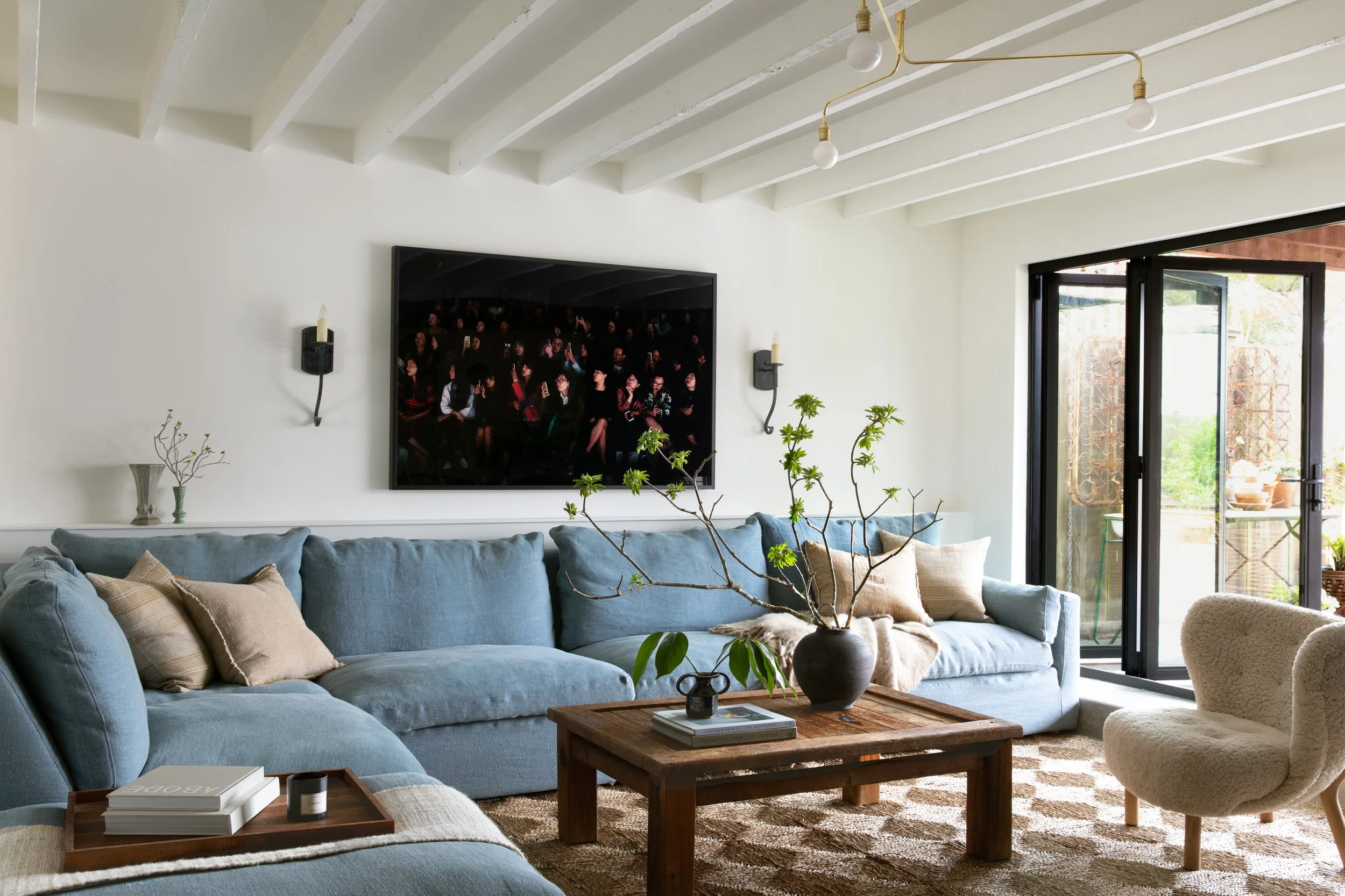 Living room view with sofa, coffee table, side chair, jute rug, vase with large branches, artwork on the wall and two wall sconces.
