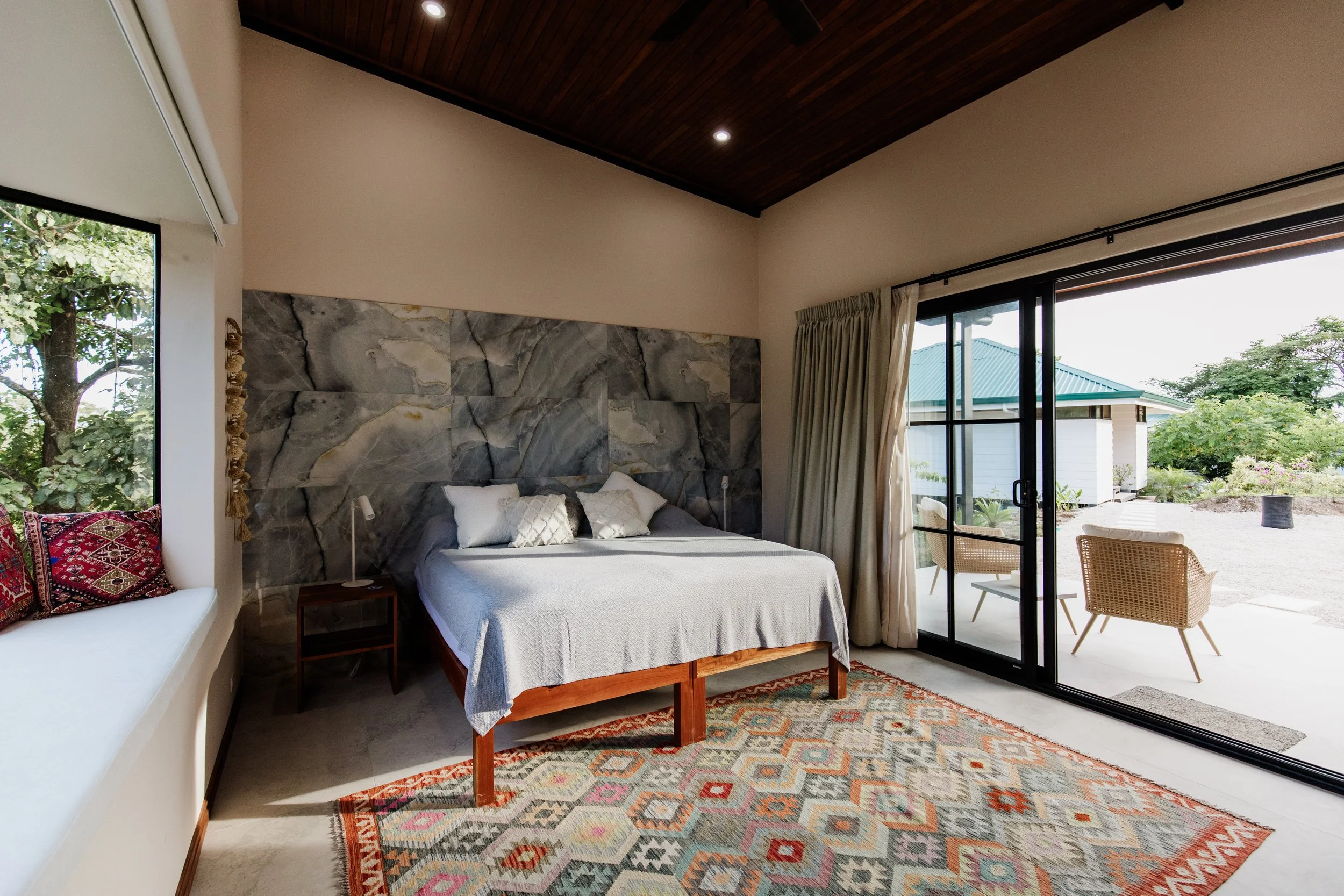 A bedroom with a wooden ceiling, marble accent wall behind the bed, and sliding glass doors leading to an outdoor patio with chairs and greenery.