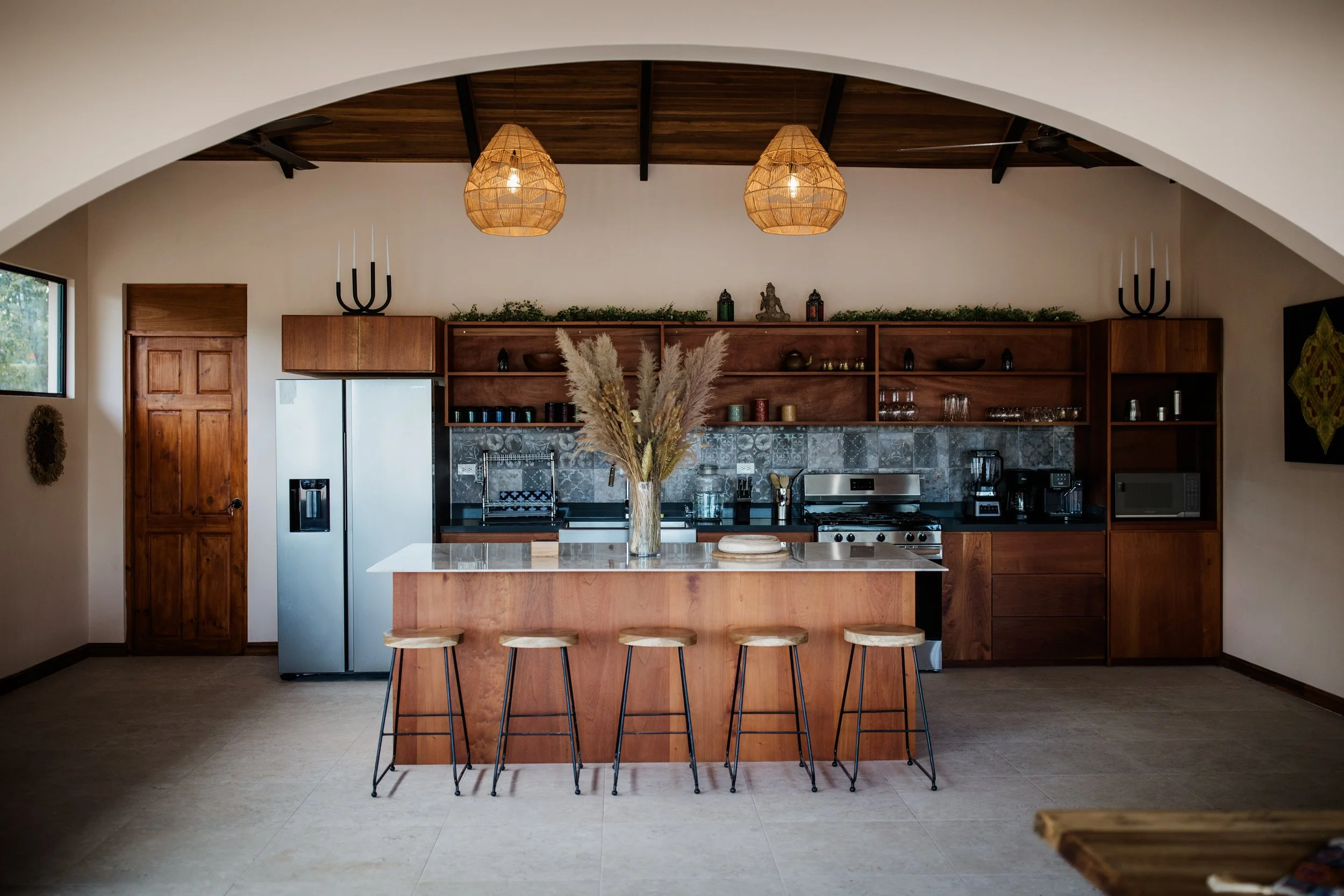 Modern kitchen with wooden cabinets, a large island with four stools, a vase with pampas grass, and pendant lights hanging from a wooden ceiling.