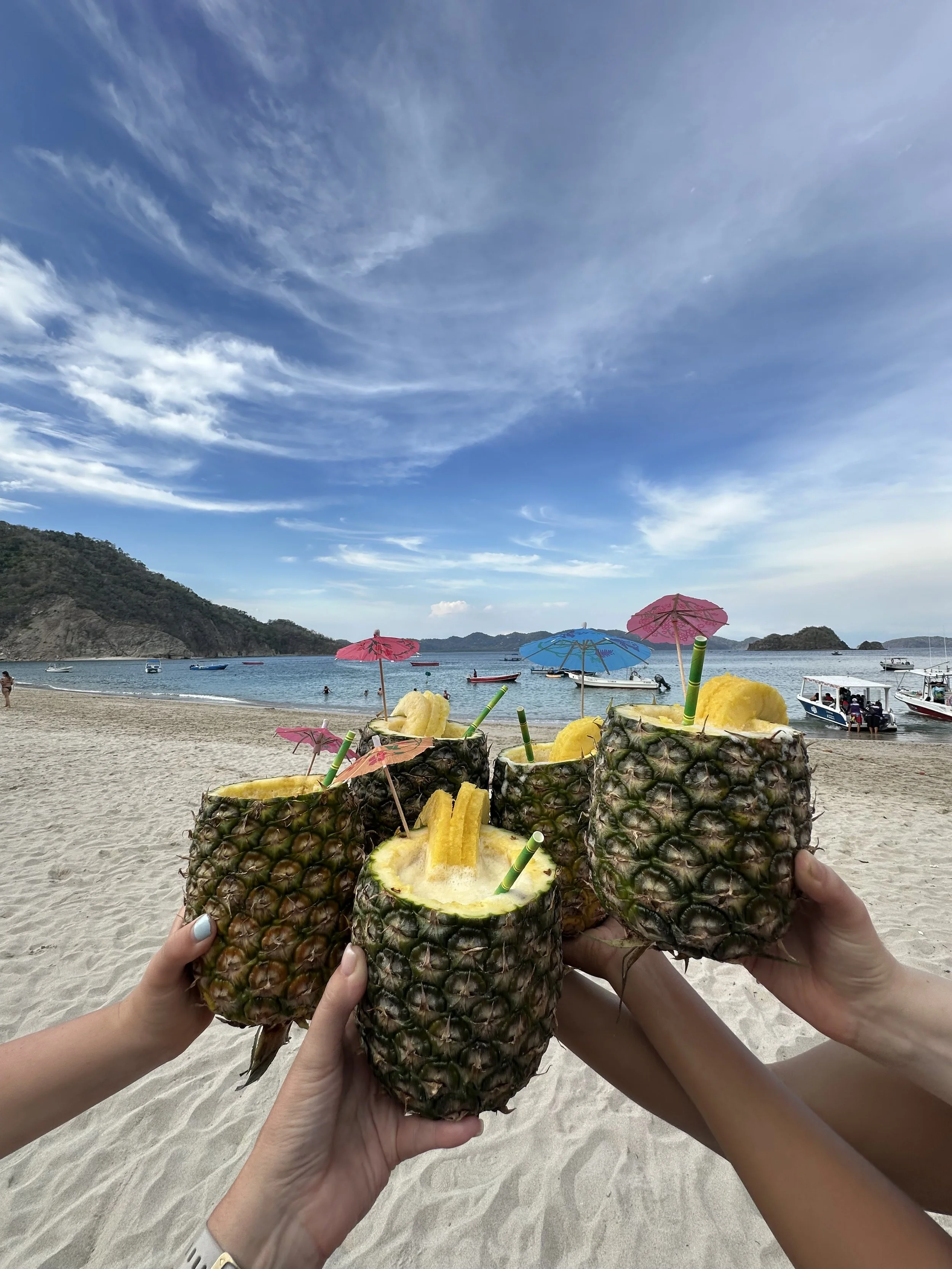 Hands holding five hollowed-out pineapples with pineapple chunks, paper umbrellas, and straws, on a sandy beach with boats on the water and a cloudy blue sky in the background.