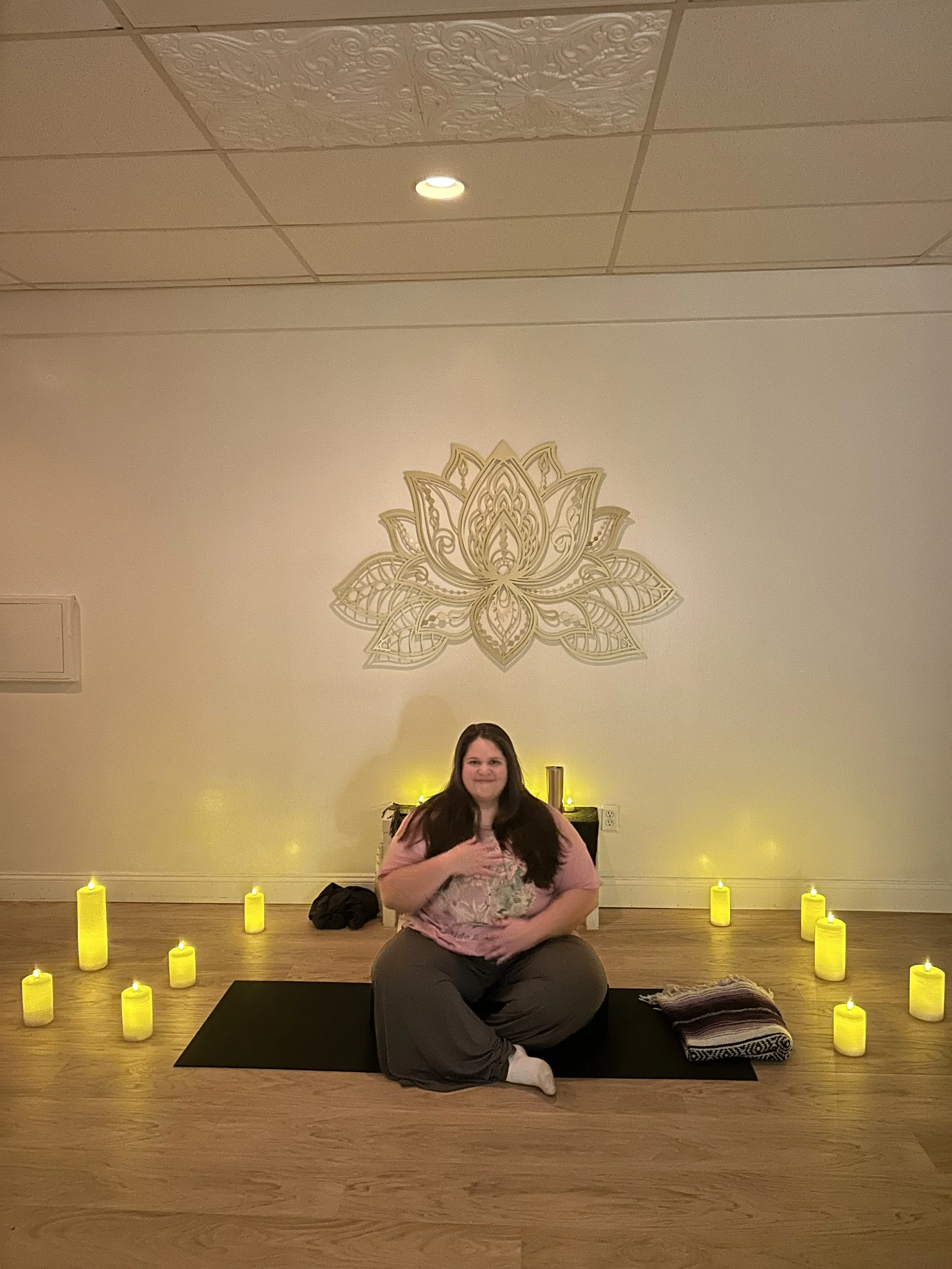 A woman sitting on a yoga mat in a meditation room with yellow candles surrounding her, a blanket to her side, and a decorative wall art of a lotus flower behind her.