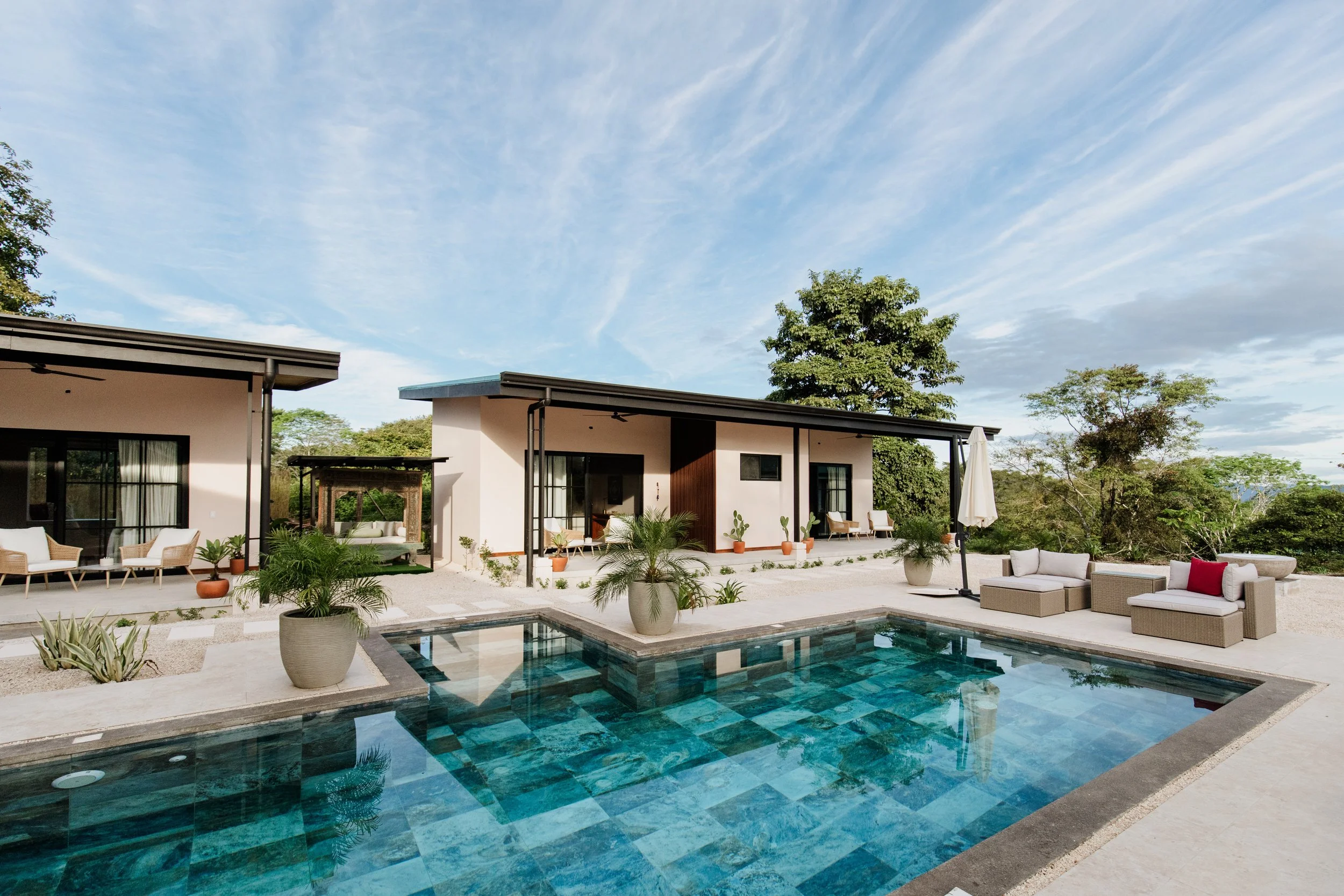 Modern outdoor patio with a swimming pool, lounge chairs, potted plants, and a house with large windows and a covered porch, surrounded by trees under a partly cloudy sky.