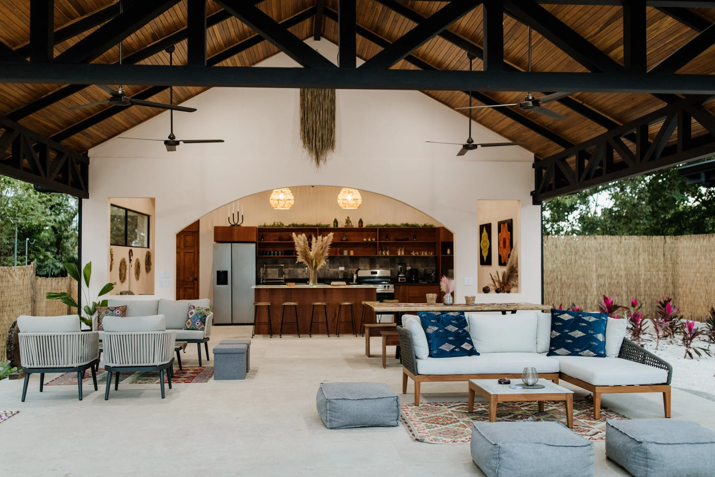 Spacious indoor living area with white sofas, blue patterned cushions, and two gray poufs, open kitchen in the background with bar stools, wooden accents, and decorative plants, ceiling fans, and a high vaulted wooden ceiling.