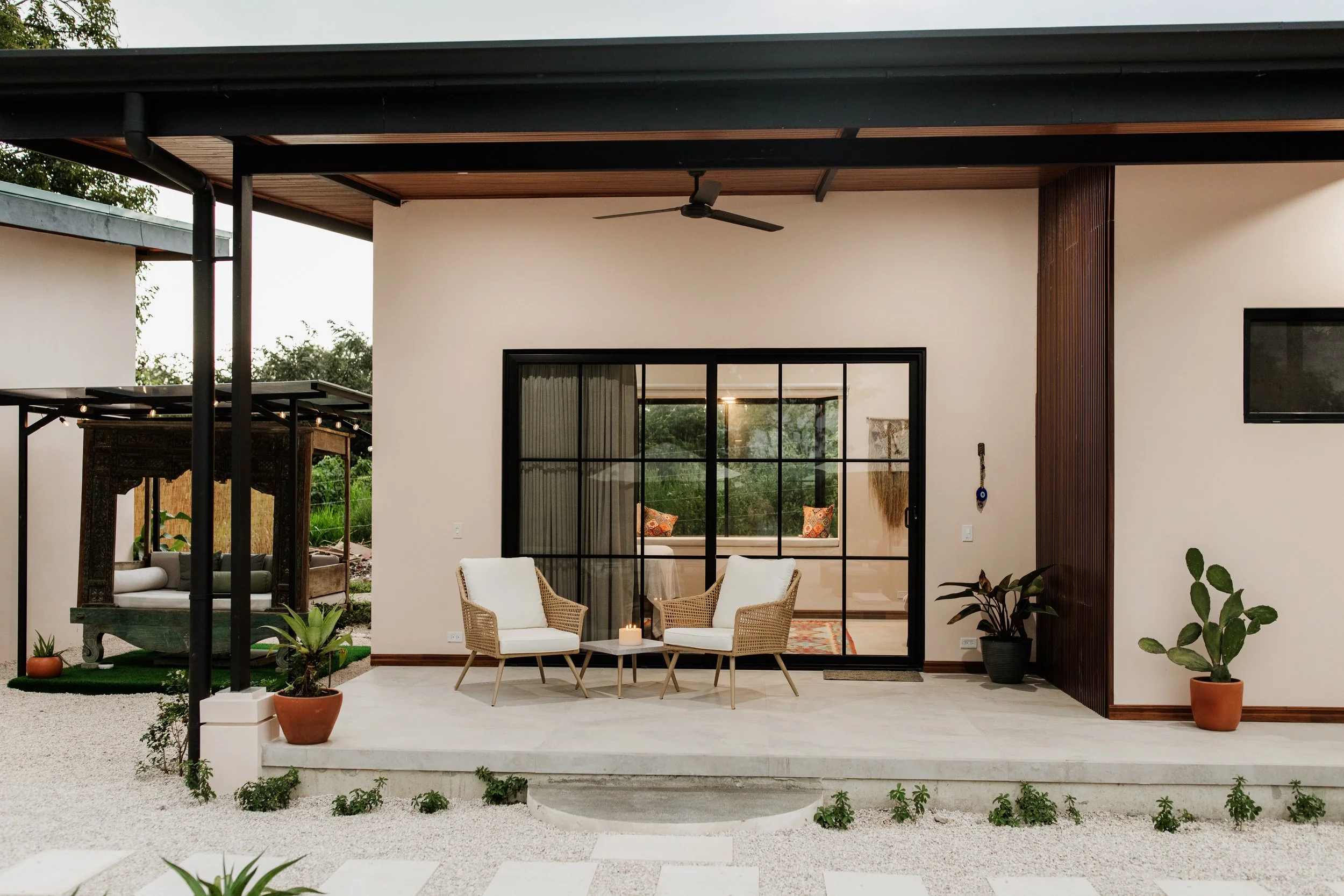 Modern house outdoor patio with two white chairs, small table with candle, potted plants, glass sliding doors, and a ceiling fan
