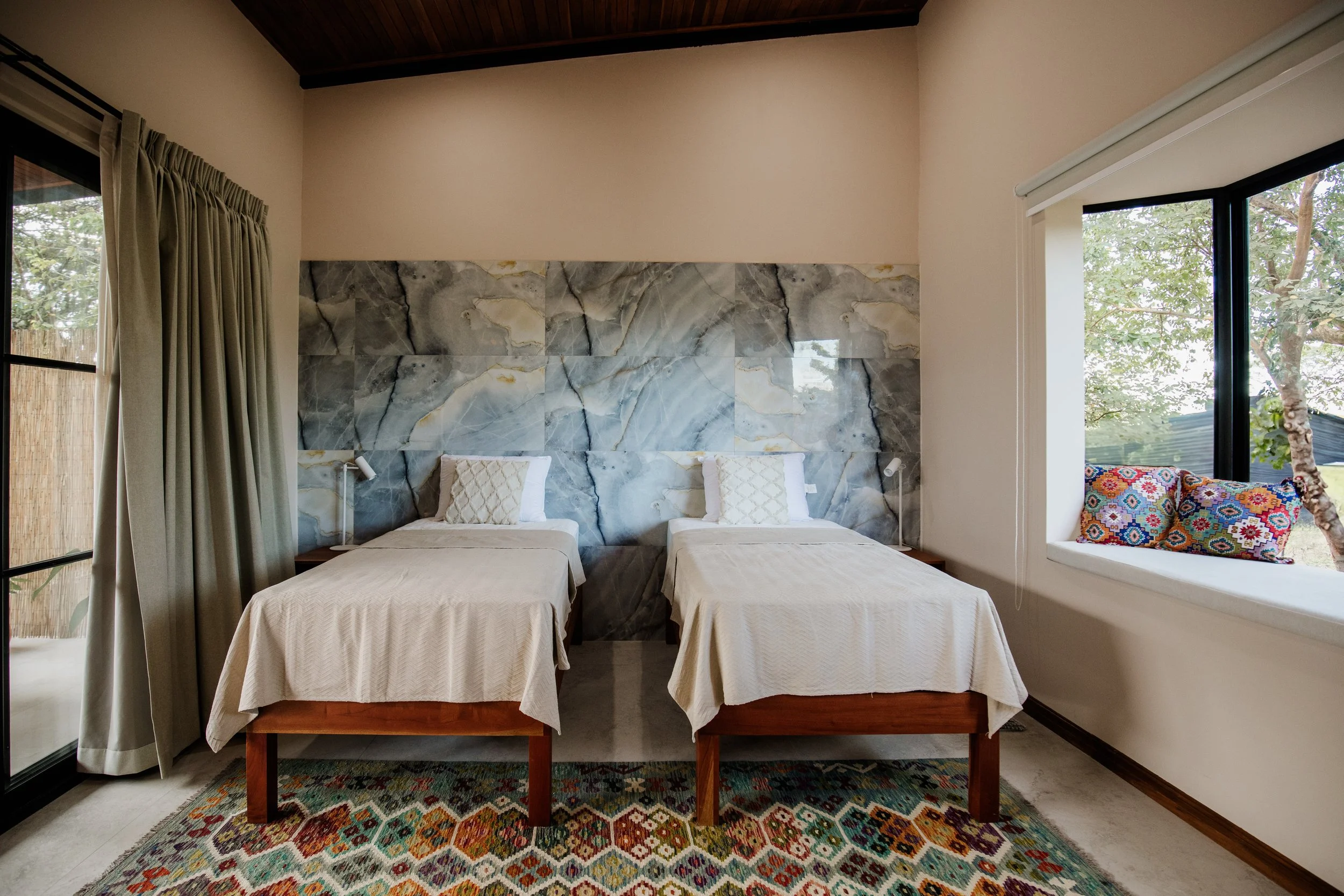 A bedroom with two single beds with white bedding, pillows, a marble accent wall, large window with curtains, and a window seat with colorful cushions.