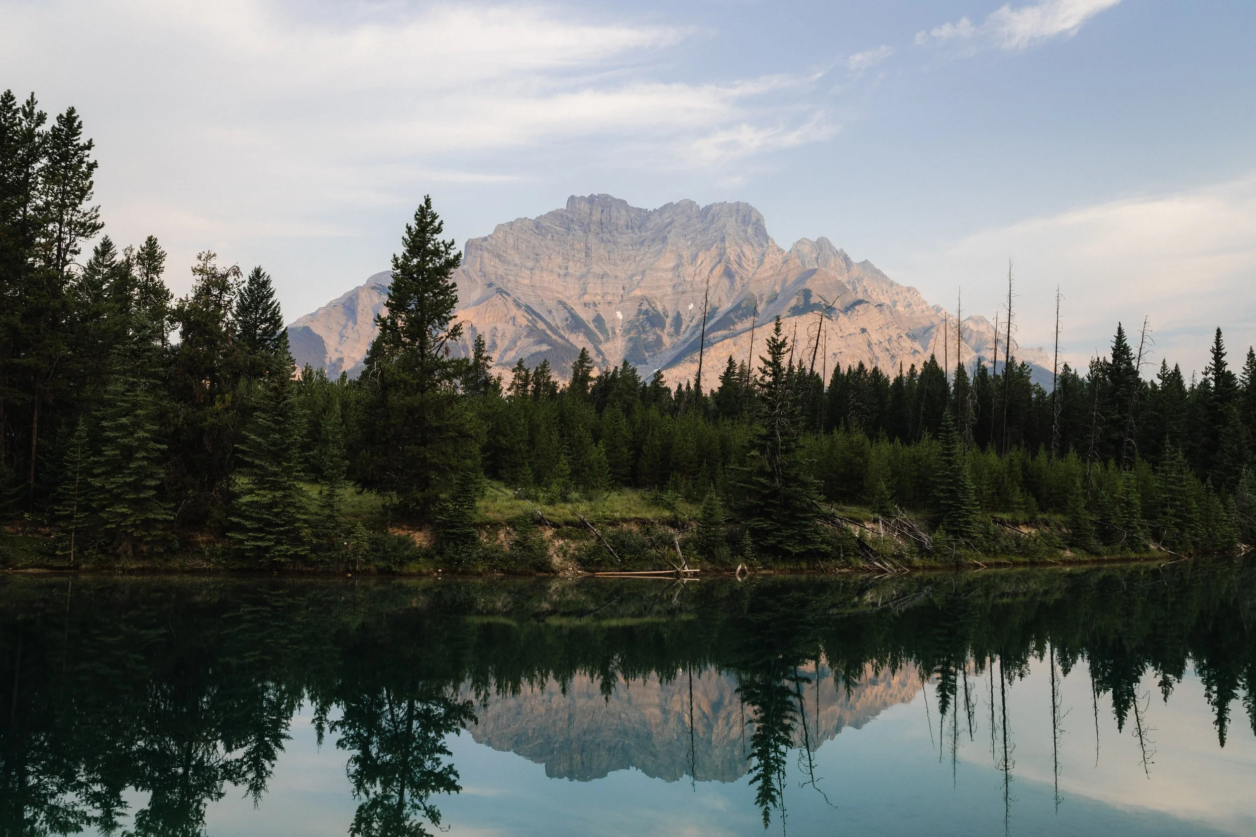 Mountain range with forest and reflection on a lake under a partly cloudy sky.