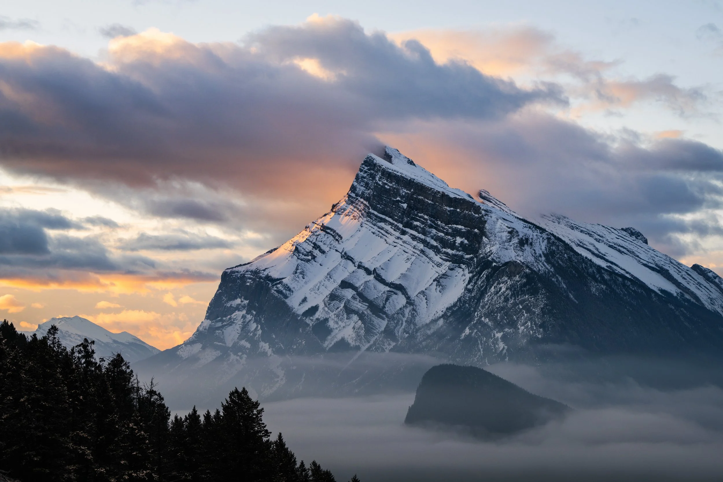 Snow-covered mountain peak with clouds and fog at sunrise or sunset.