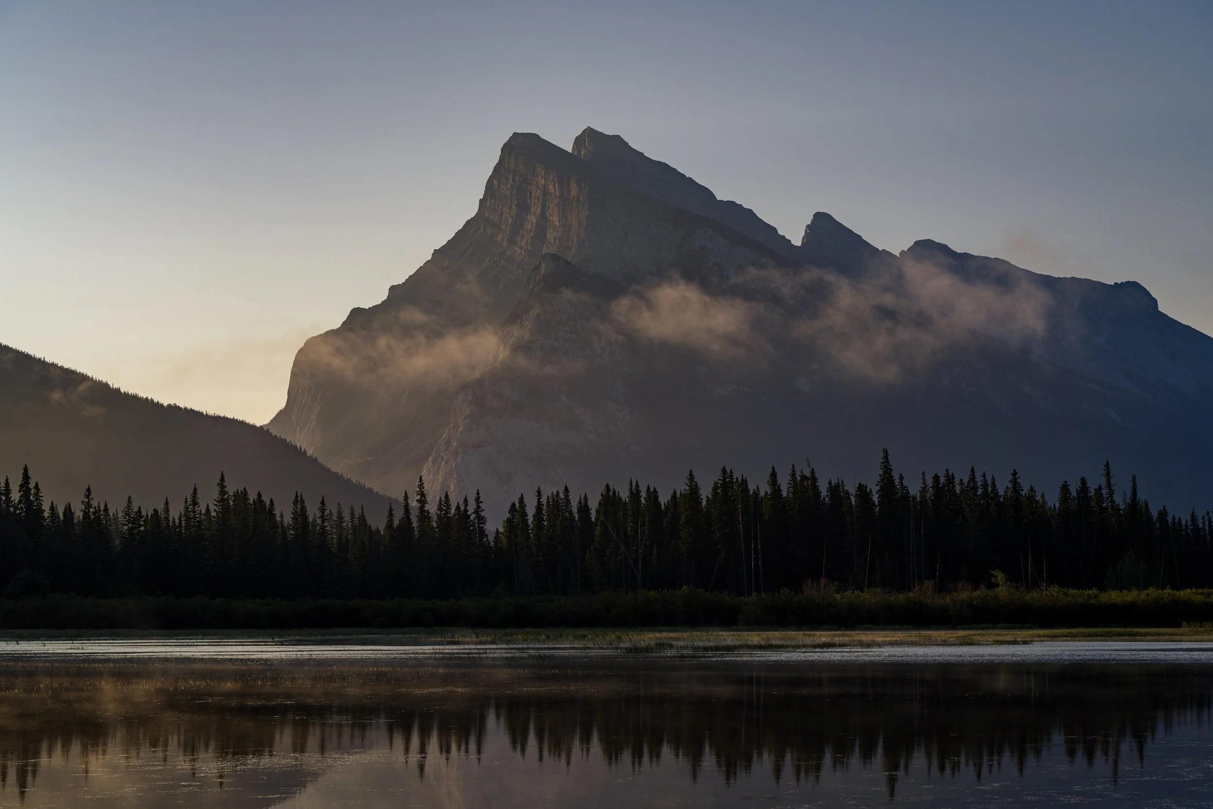 A mountain with a forest at the base and a calm body of water reflecting the scenery in the foreground, under a clear sky.