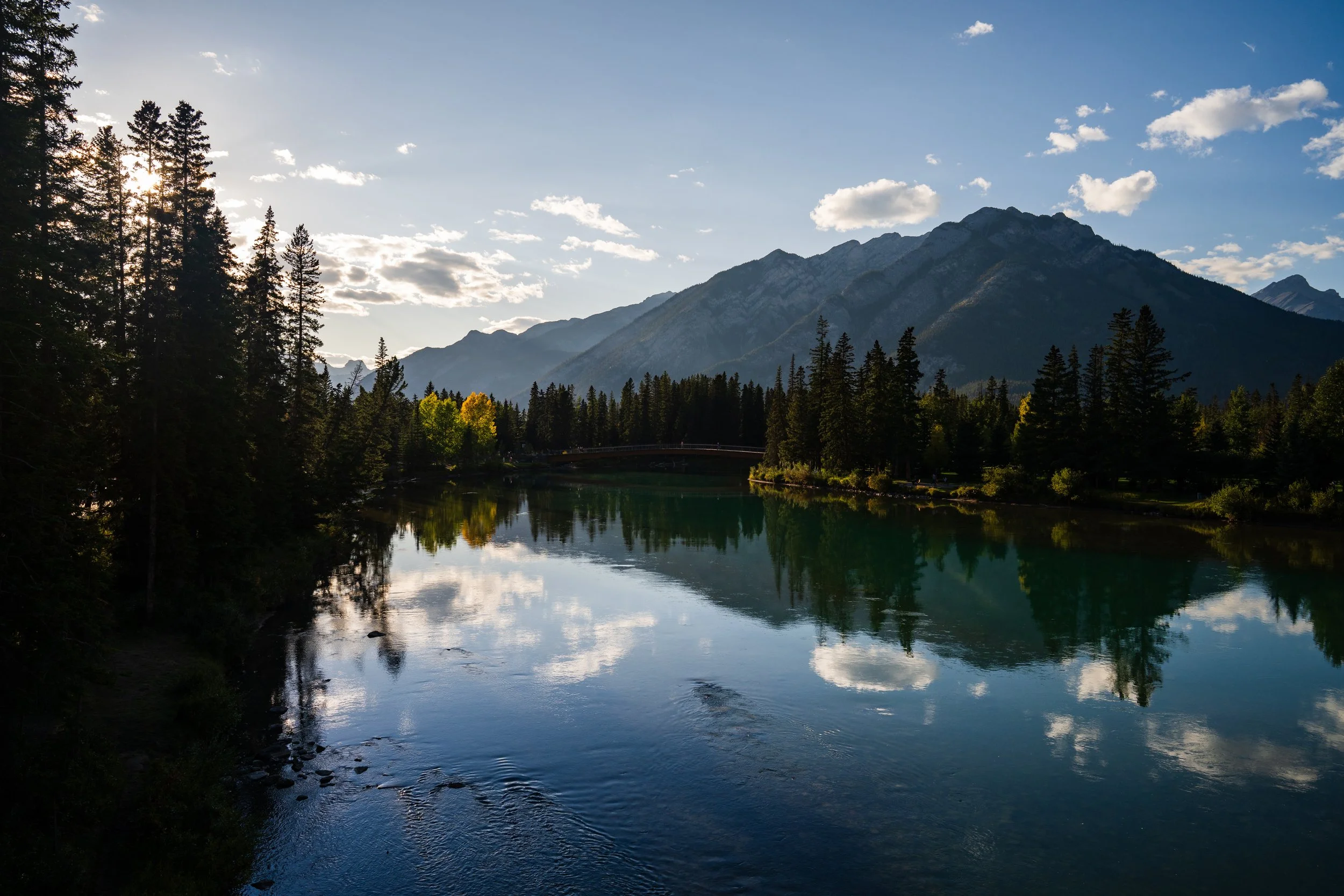 A scenic view of a lake surrounded by tall pine trees, with a backdrop of mountain peaks and a partly cloudy sky reflected in the water.