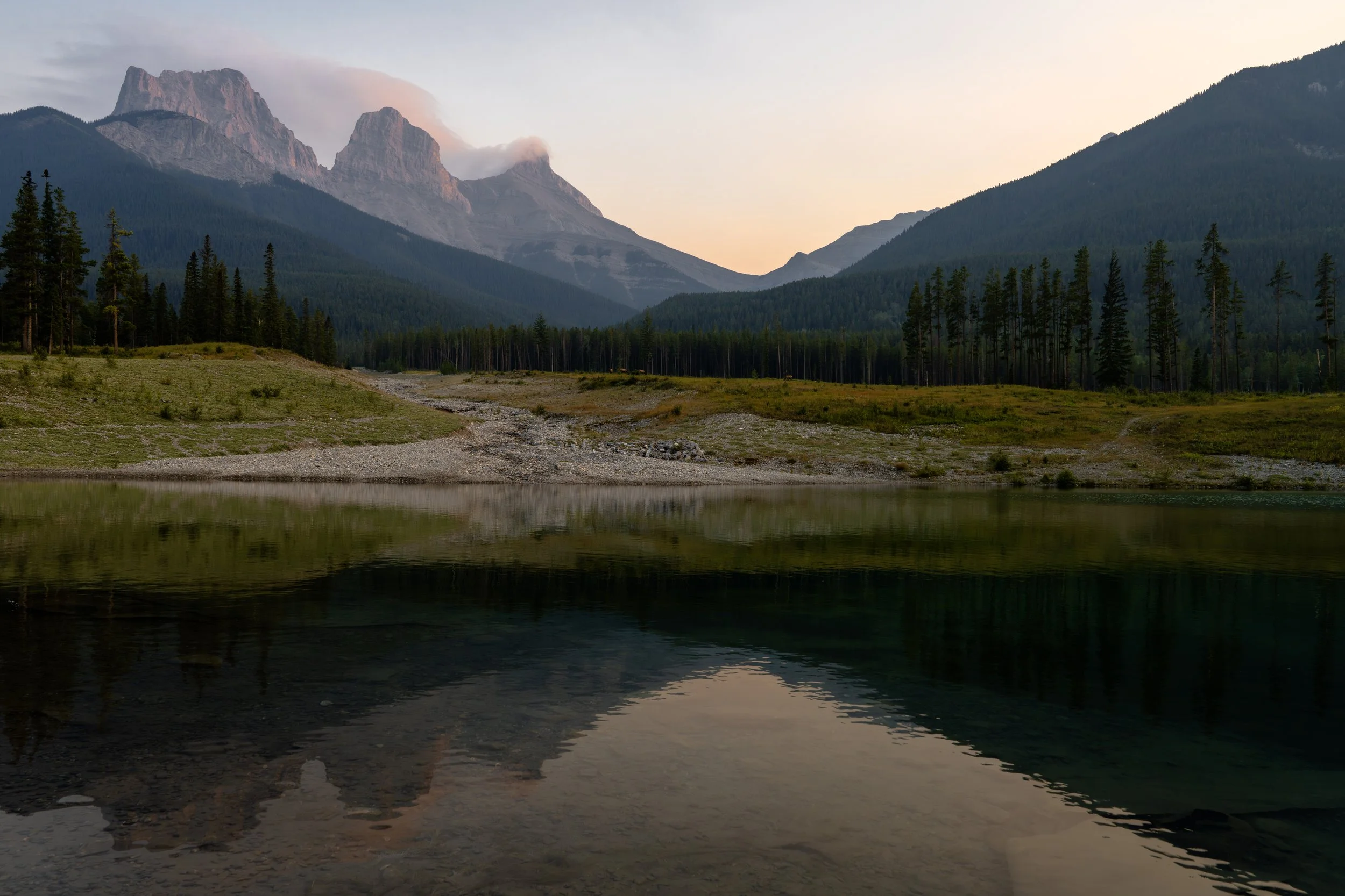 A serene mountain lake scene at dusk, with calm water reflecting the surrounding trees and distant mountains.
