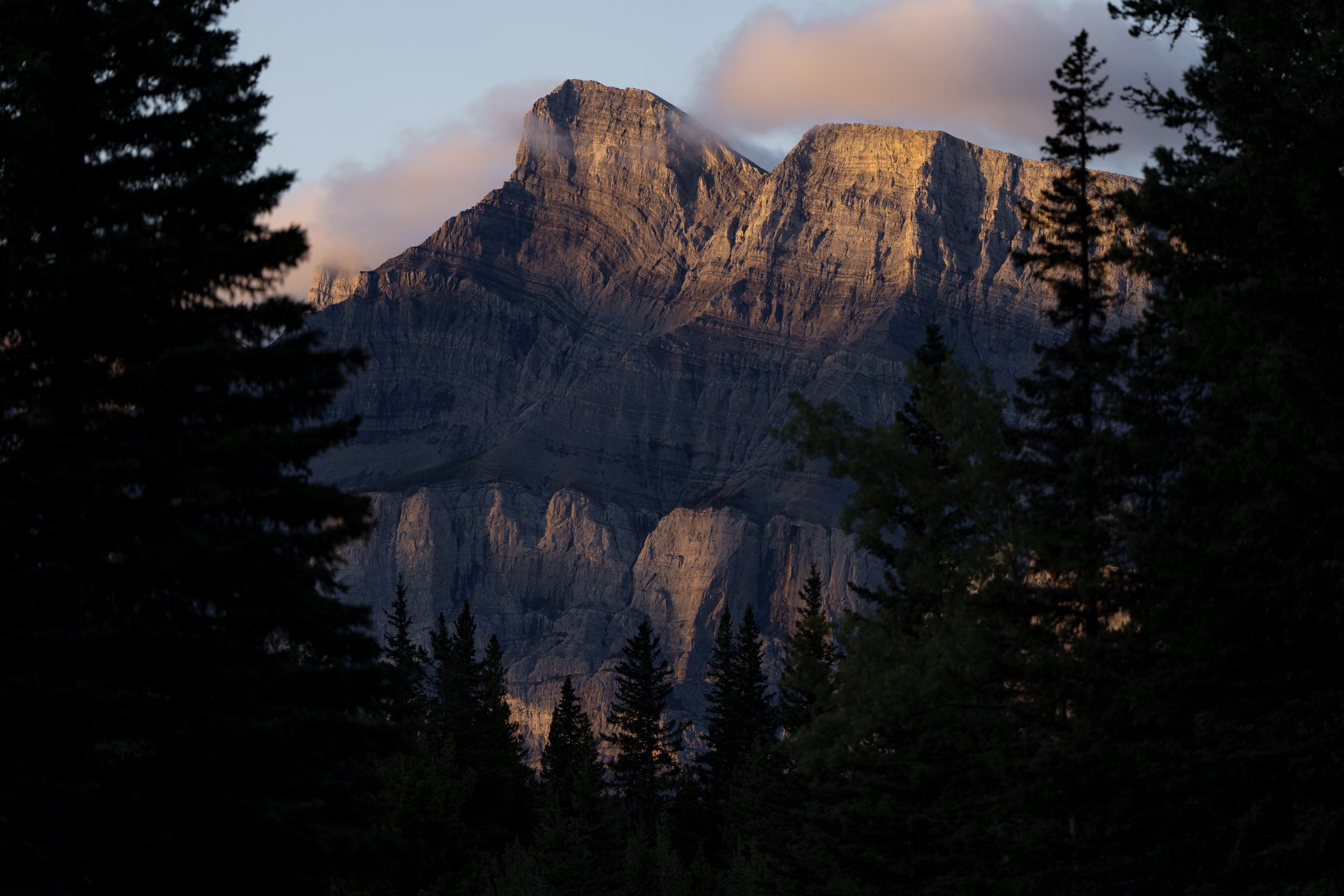 A mountain landscape during sunset with a towering mountain peak surrounded by trees in the foreground.