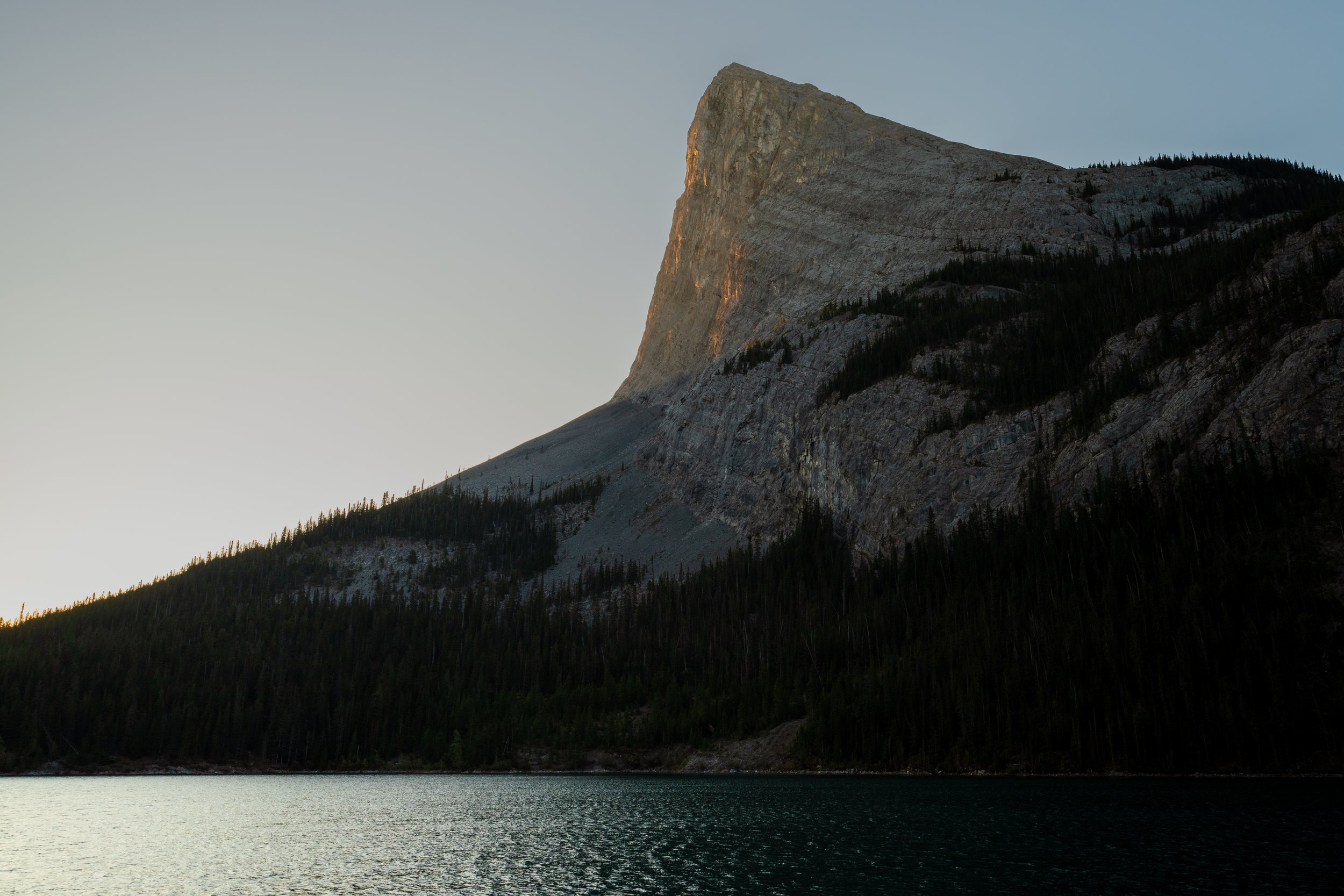 A large mountain with a prominent peak rises above a forested hillside and a body of water in the foreground. The scene appears to be at dusk or dawn.
