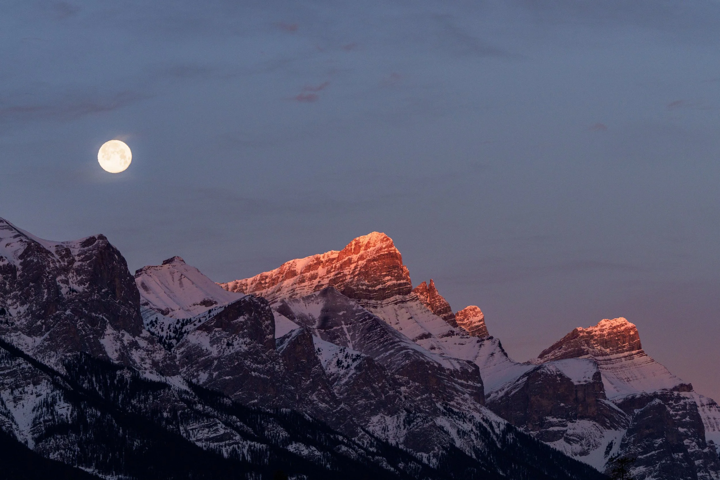 Nighttime mountain landscape with snow-capped peaks illuminated by the rising sun, under a sky with a full moon and scattered clouds.